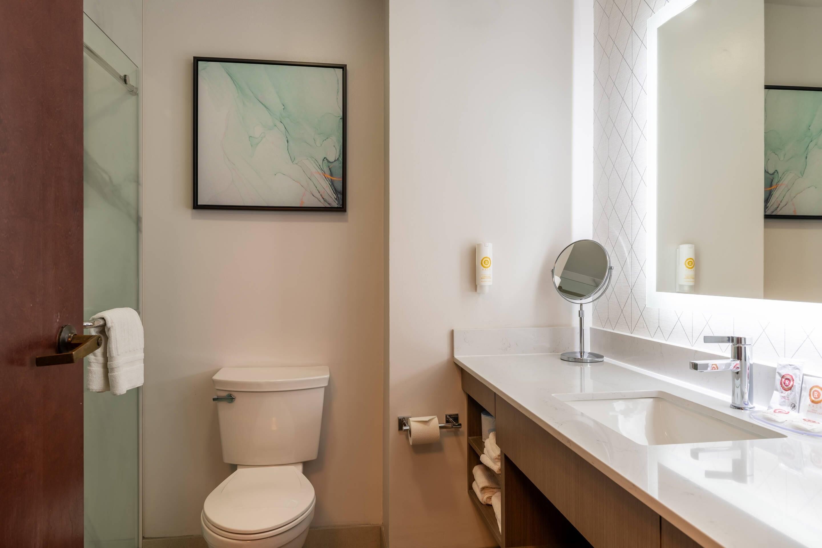 A modern bathroom with a toilet, a sink with a mirror, and a framed abstract artwork on the wall.