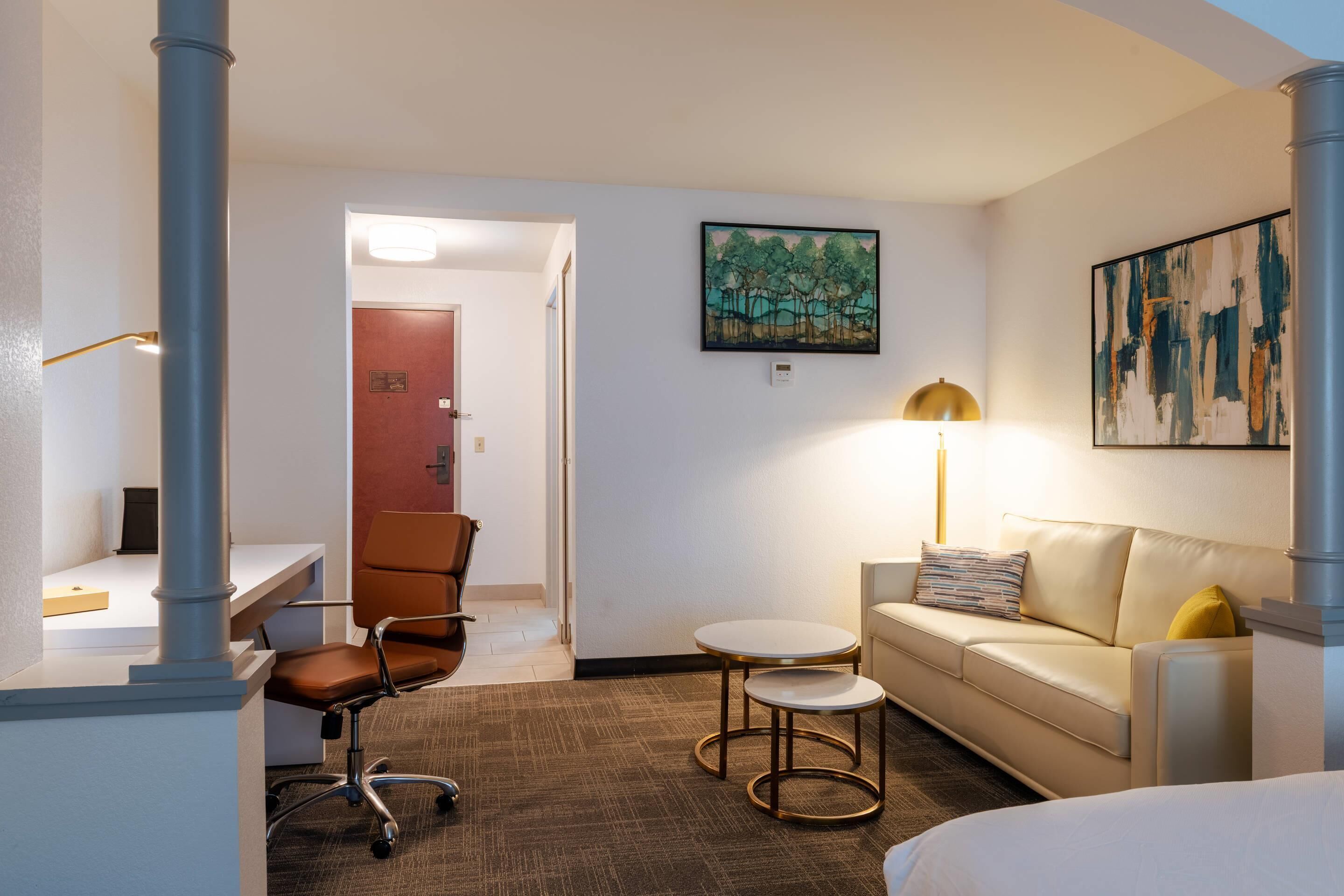 A hotel room with a desk, office chair, sofa, and a small round table. There is a painting on the wall and a lamp on the sofa.