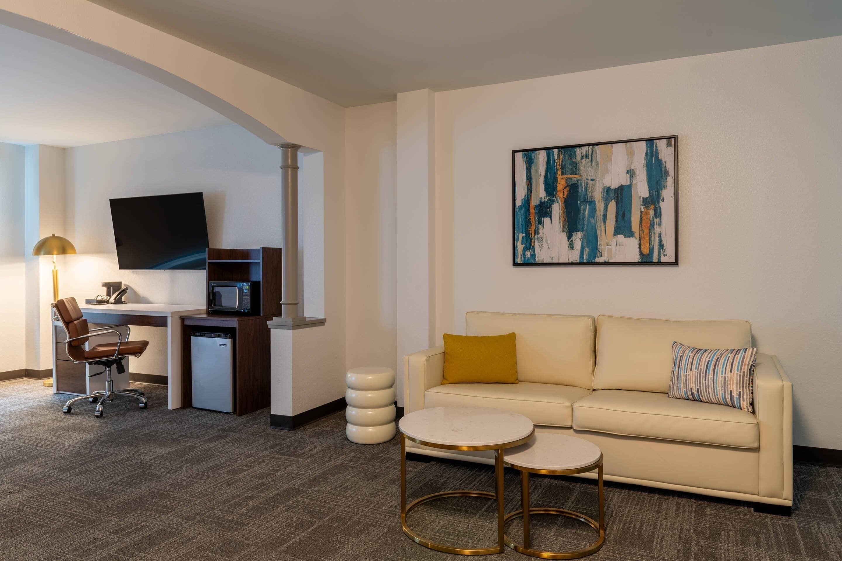 Een hotelkamer met een bank, twee kleine ronde tafels, een bureau met een computer en een televisie, en een modern abstract schilderij aan de muur.