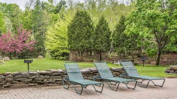 Un patio con tres sillones verdes, un muro de piedra y una zona verde con árboles, incluyendo un árbol de flores rosadas.