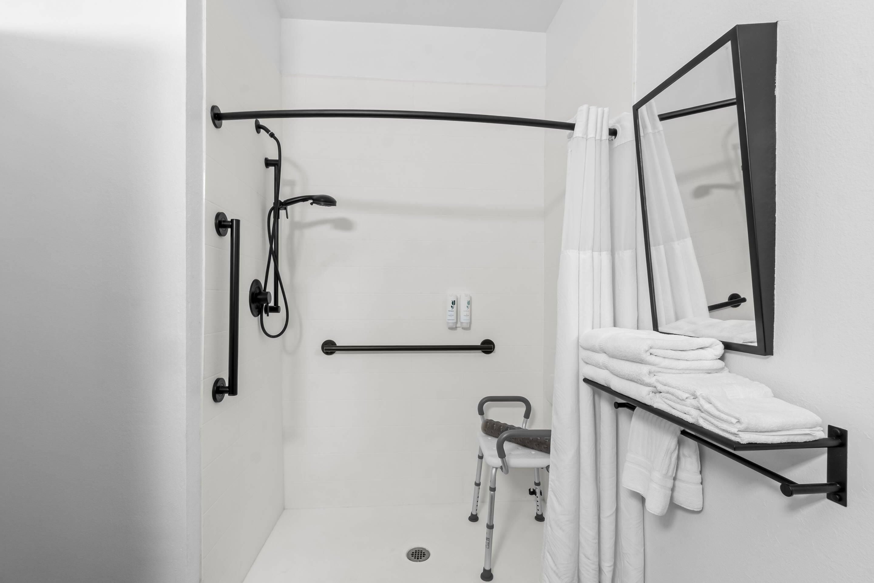 Une salle de bain blanche équipée d’une douche, d’une barre d’appui, d’une poignée de maintien, d’un fauteuil roulant et d’un porte-serviettes.