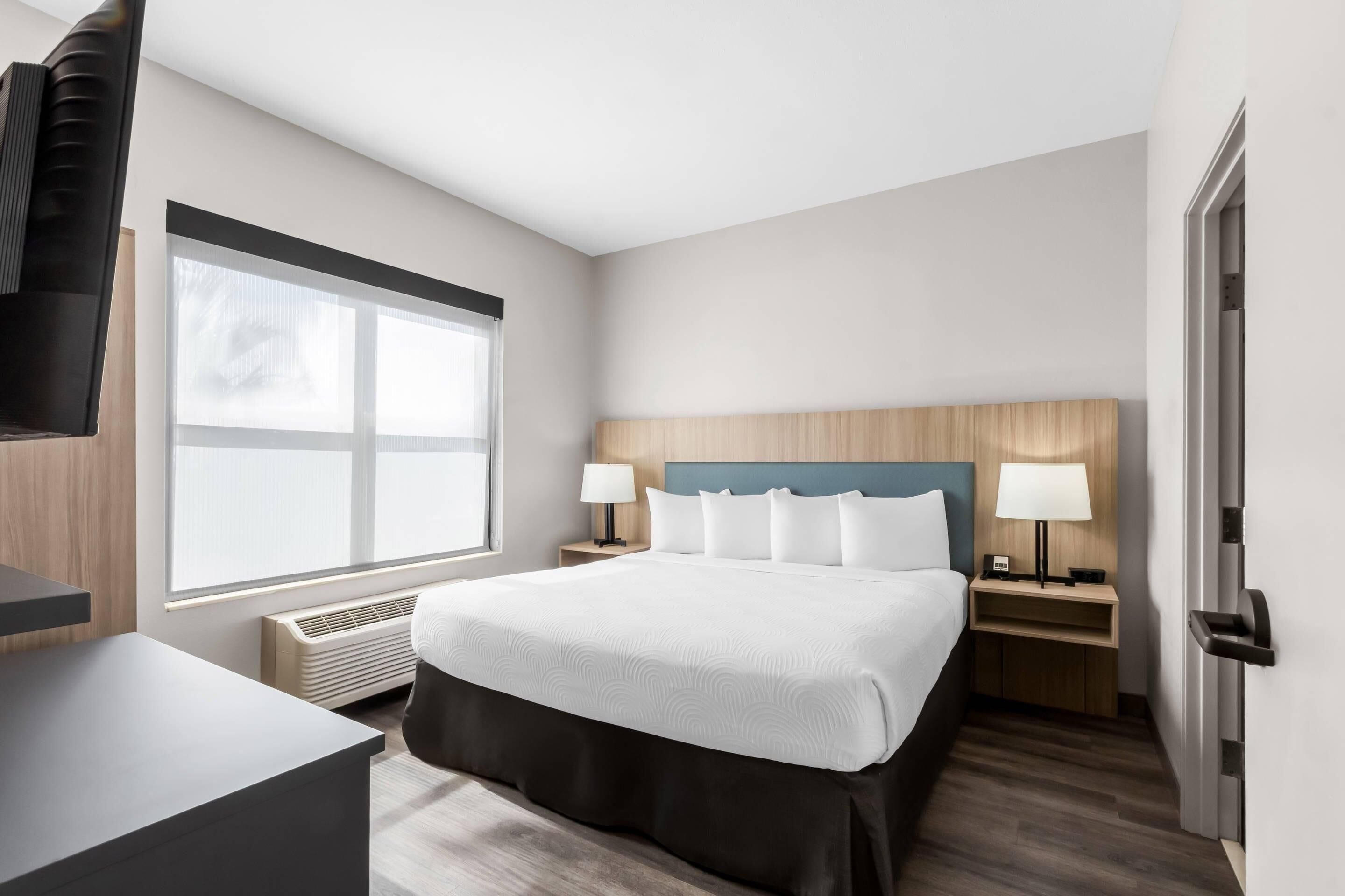 A hotel room with a bed, bedside tables, a desk, a window, and a door.