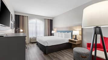 A hotel room with a bed, bedside tables, a desk, a TV, a red chair, a lamp, and a window with curtains.