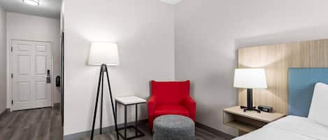 Habitación de hotel con una cama, una silla roja, una mesita con lámpara y una mesita de noche con lámpara y reloj.