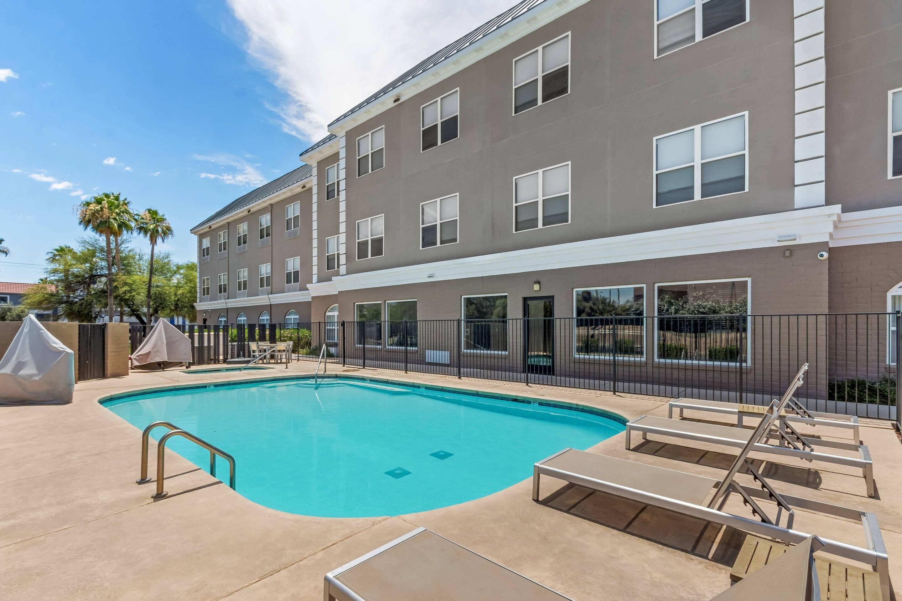 A swimming pool with lounge chairs, a covered seating area, and a multi-story building with large windows.