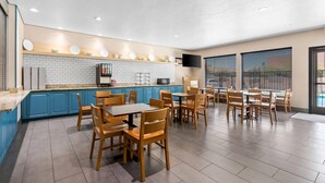 A spacious dining area with blue cabinets, wooden tables and chairs, a kitchen area with white tiles, and a flat-screen TV mounted on the wall. - Country Inn & Suites by Radisson, Tucson Airport, AZ (Tucson)