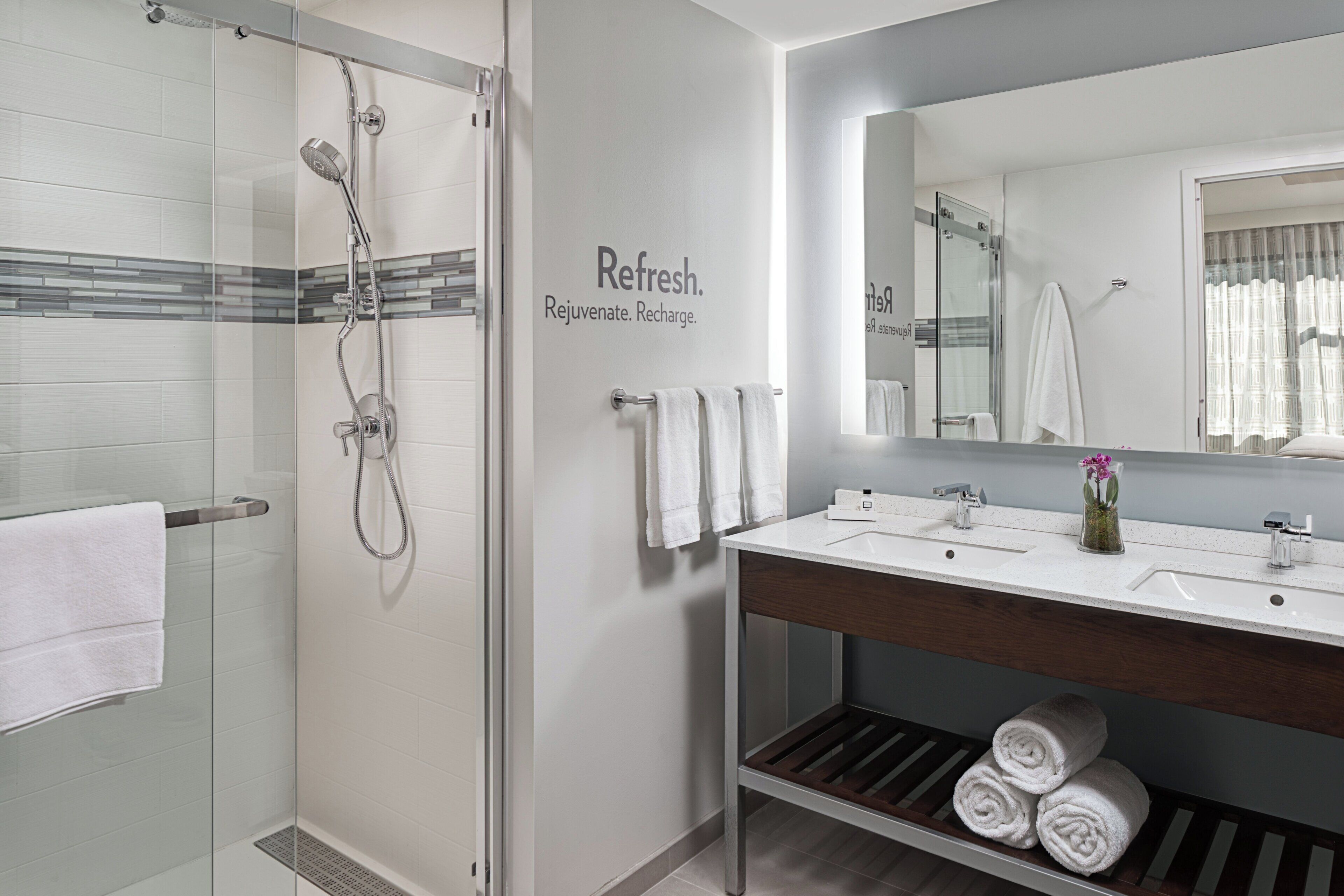 A modern bathroom with a glass shower enclosure, a vanity with a mirror, and a shelf with rolled towels.