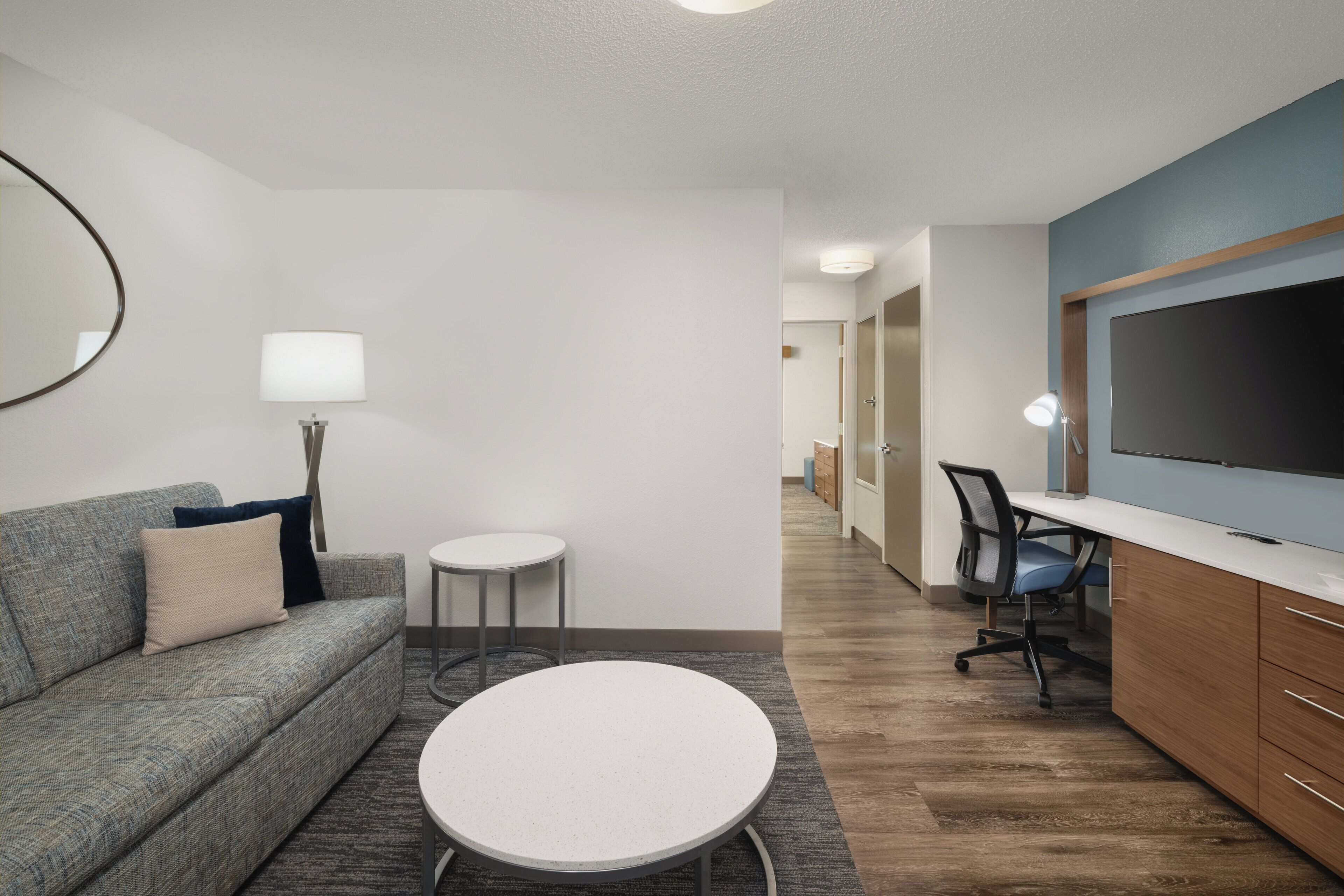 Una habitación de hotel moderna con un sofá gris, una mesa redonda blanca, un escritorio con una silla, un televisor de pantalla plana y un espejo grande en la pared.