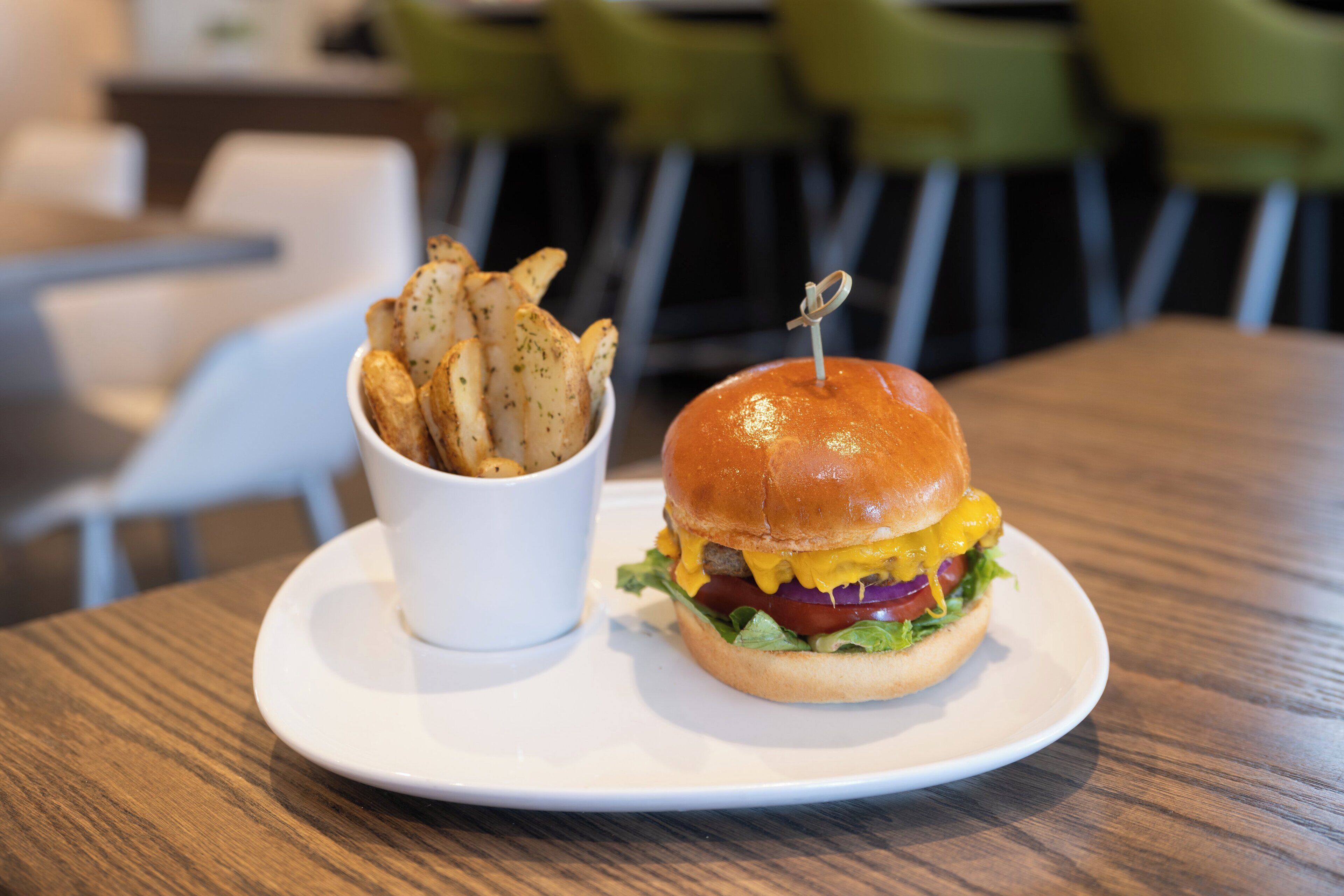 A burger with a bun, lettuce, tomato, and cheese, and a cup of fries on a white plate.