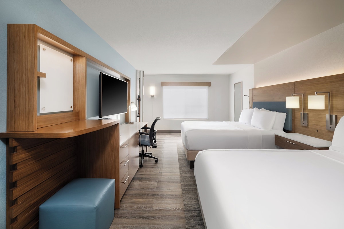 A hotel room with a wooden desk, a flat-screen TV, two beds, a desk chair, and a window.
