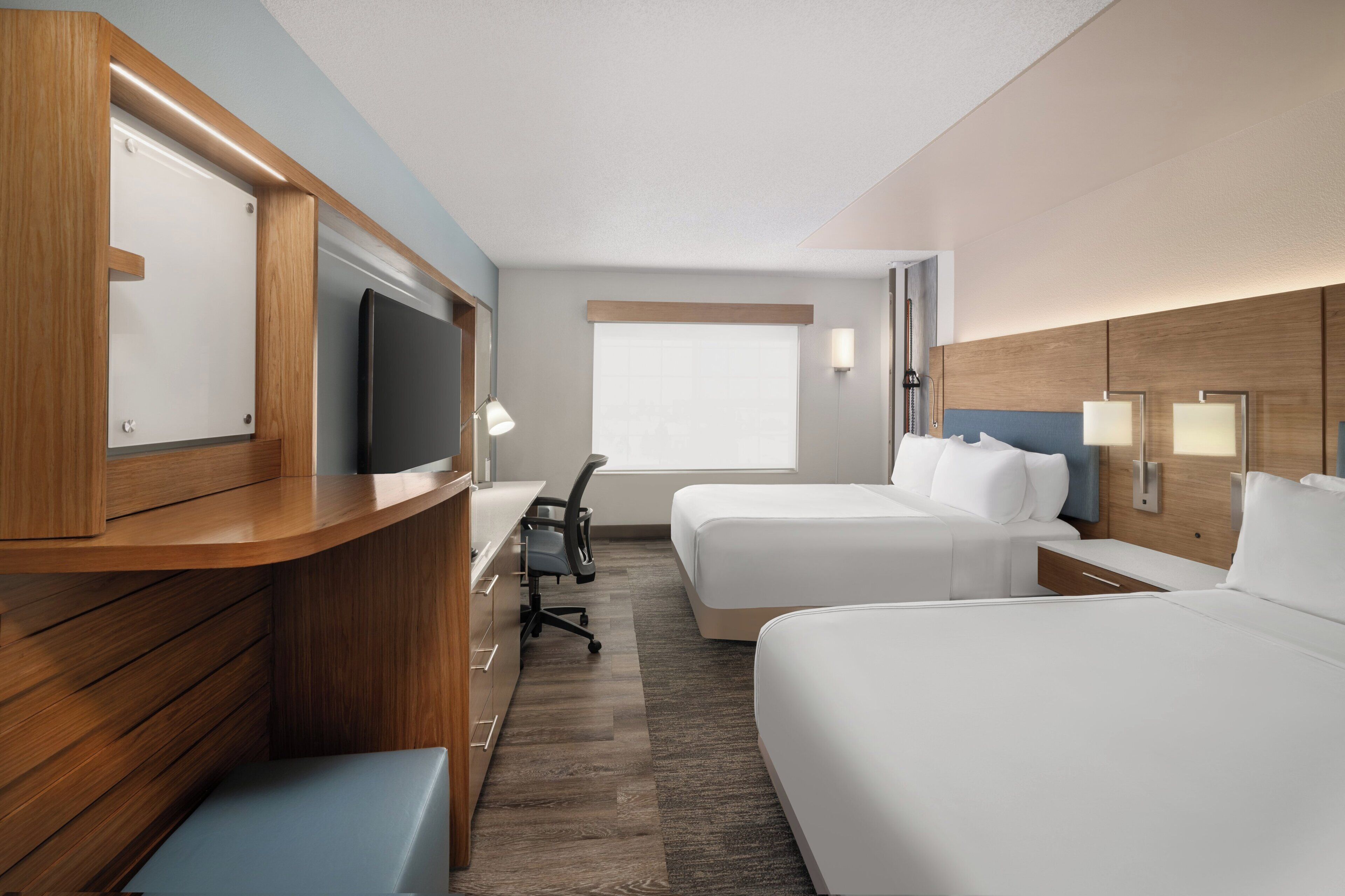 A hotel room with a wooden desk, two beds, a TV, and a window.
