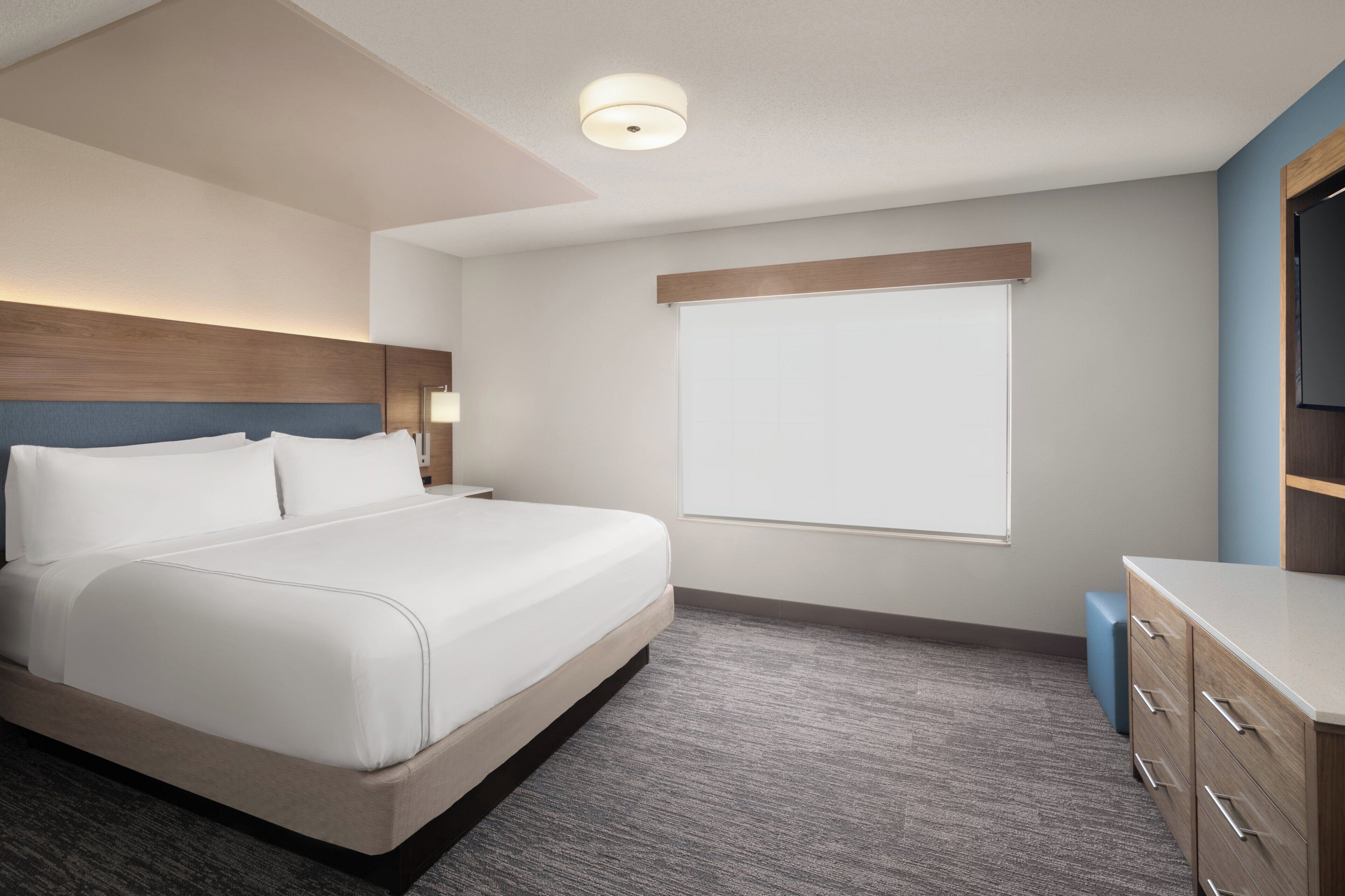 A modern hotel room with a large bed, a wooden headboard, a bedside table with a lamp, a wardrobe, and a large window.