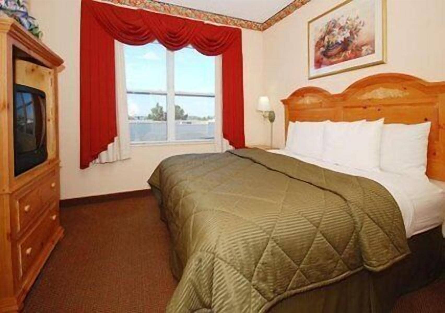 A hotel room with a bed, a wooden dresser, a window with red curtains, and a painting on the wall.