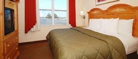 A hotel room with a bed, a wooden dresser, a window with red curtains, and a painting on the wall.