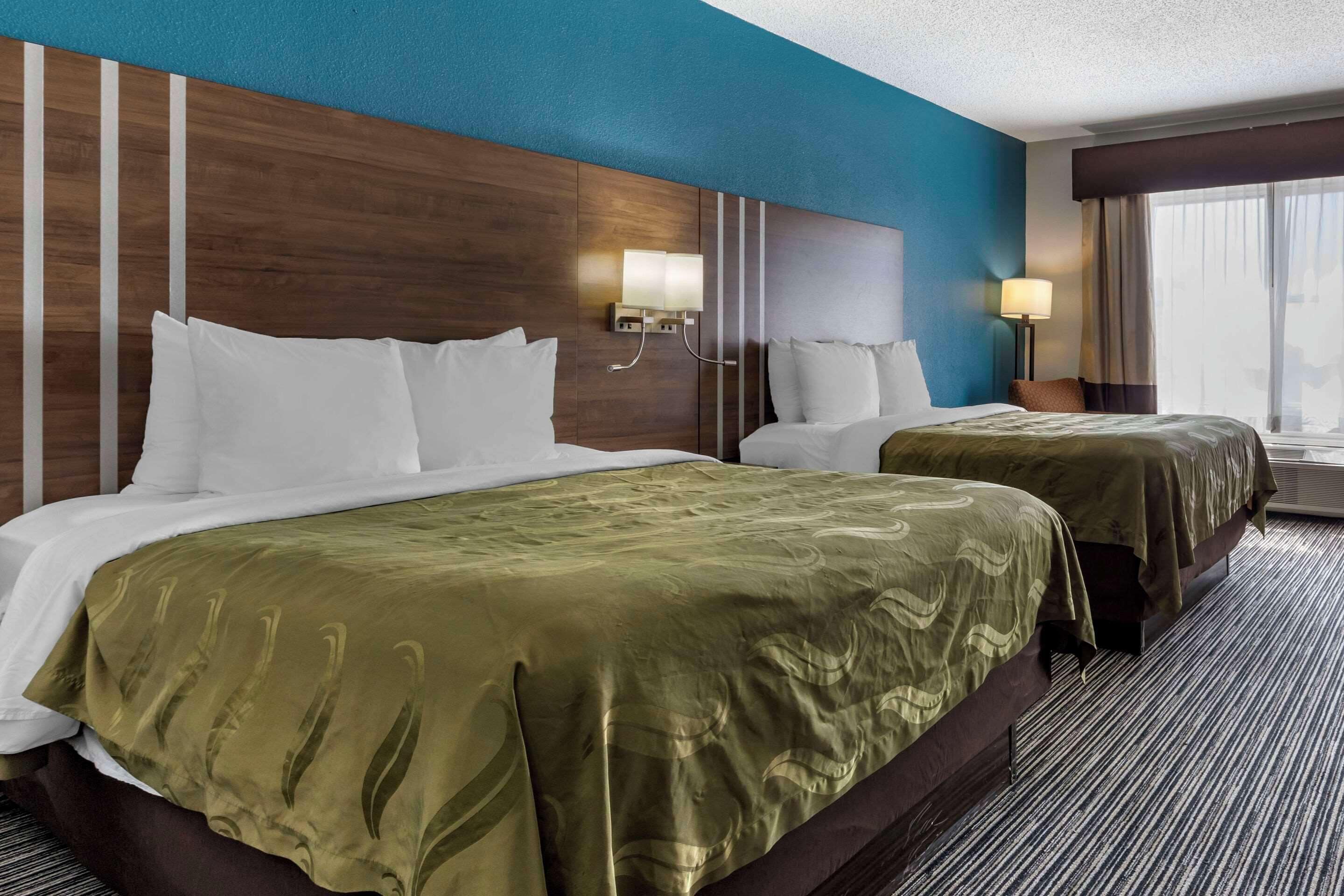 Une chambre d’hôtel avec deux lits, une tête de lit en bois, un mur d’accent bleu et une fenêtre avec des rideaux.