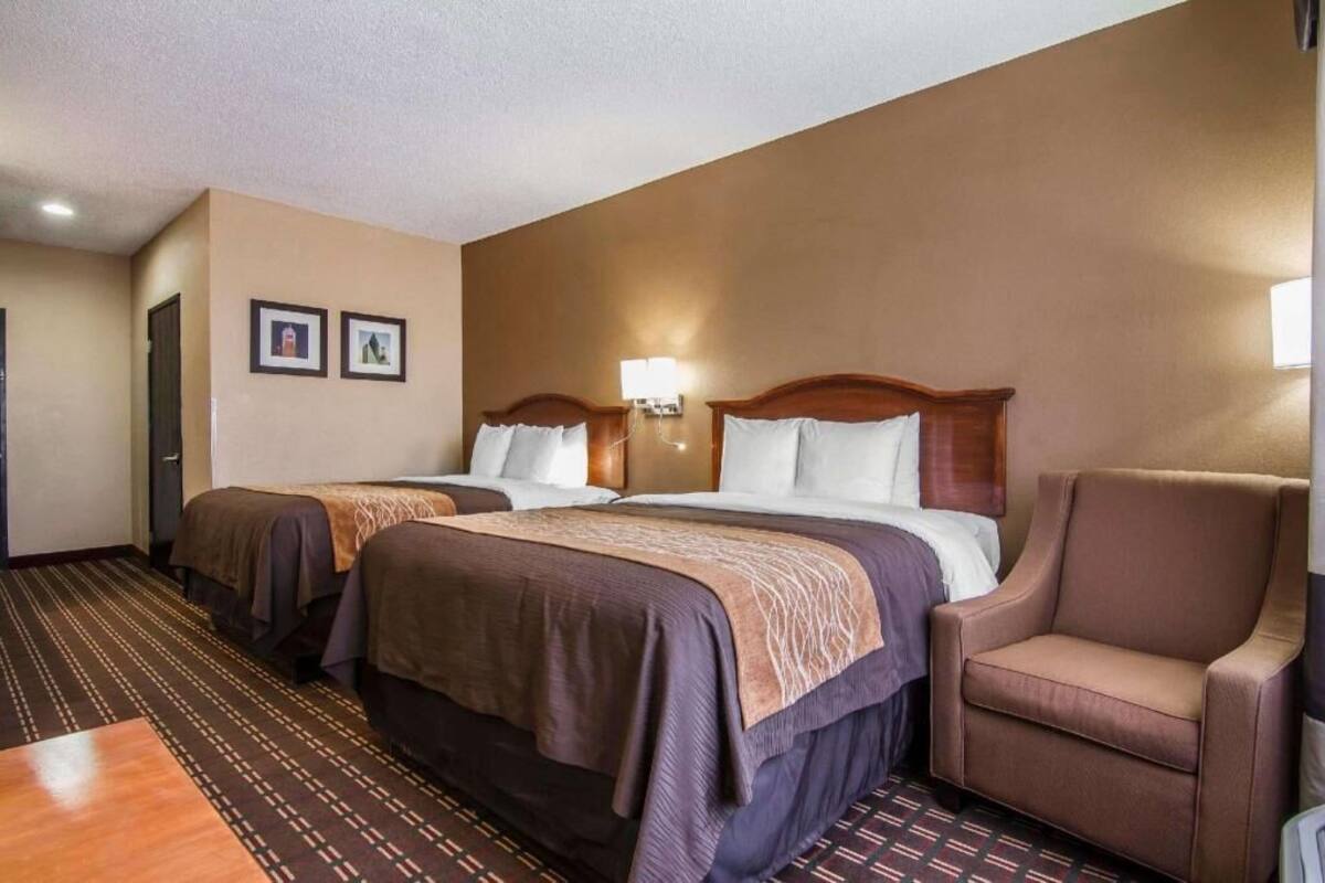 A hotel room with two beds, a chair, a carpeted floor, and a wall-mounted lamp.