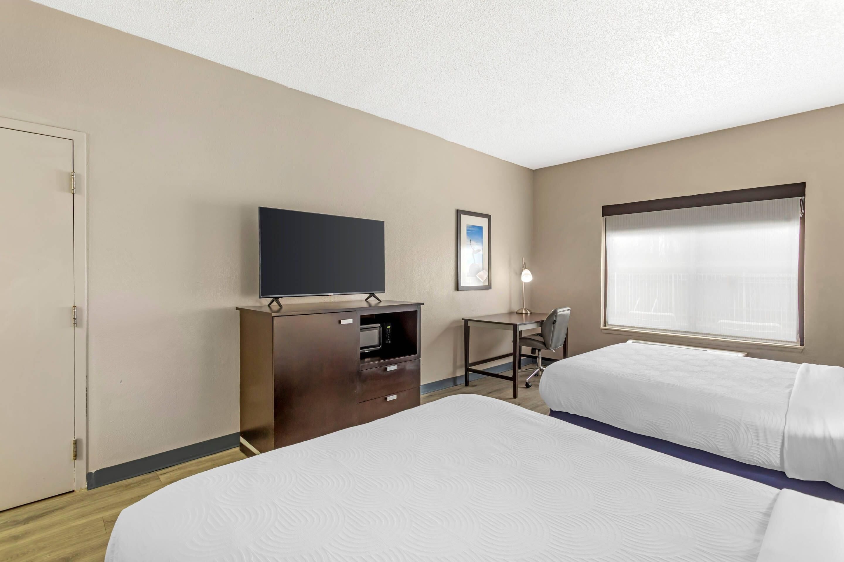 Quarto de hotel com duas camas, uma escrivaninha, televisão e janela com persianas.
