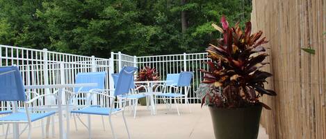 An outdoor patio area with white metal chairs and tables, surrounded by a bamboo fence, with a large potted plant in the foreground.