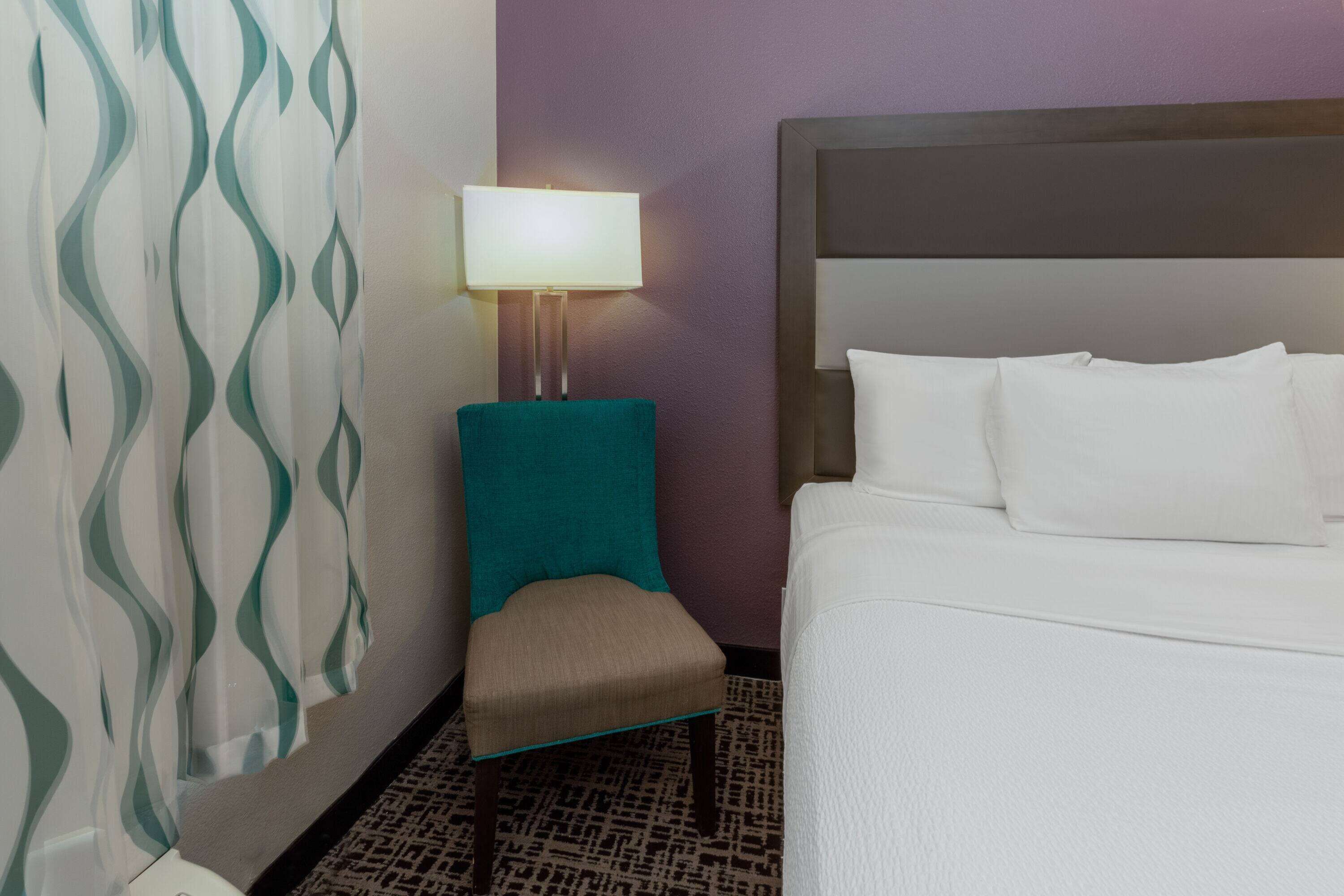 A hotel room with a bed, a teal chair, a nightstand with a lamp, and a patterned curtain.