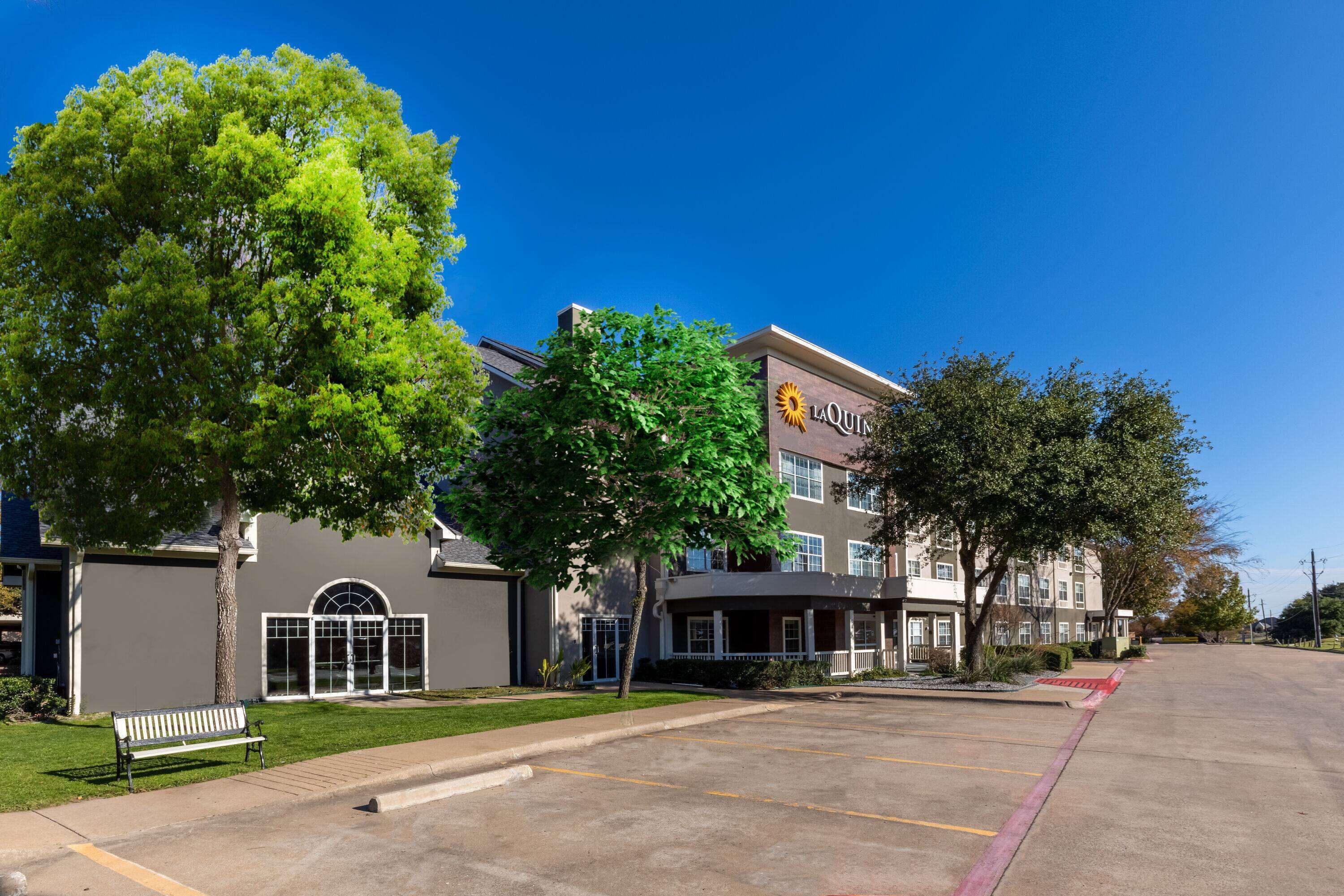 A LaQuin hotel with a parking lot, a bench, and a large tree in front.
