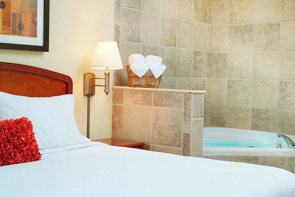 A hotel room with a bed, a nightstand, a lamp, a basket of towels, and a bathtub.