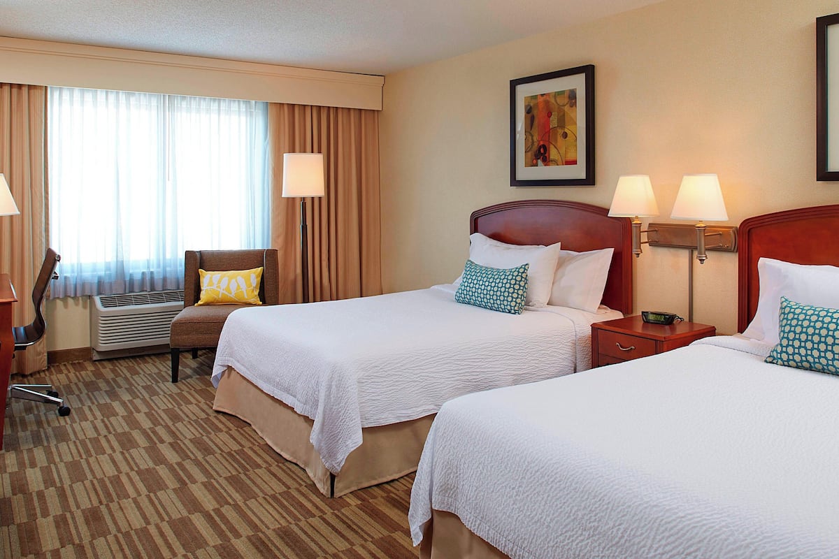 A hotel room with two beds, a chair, an air conditioning unit, and a desk with a lamp.