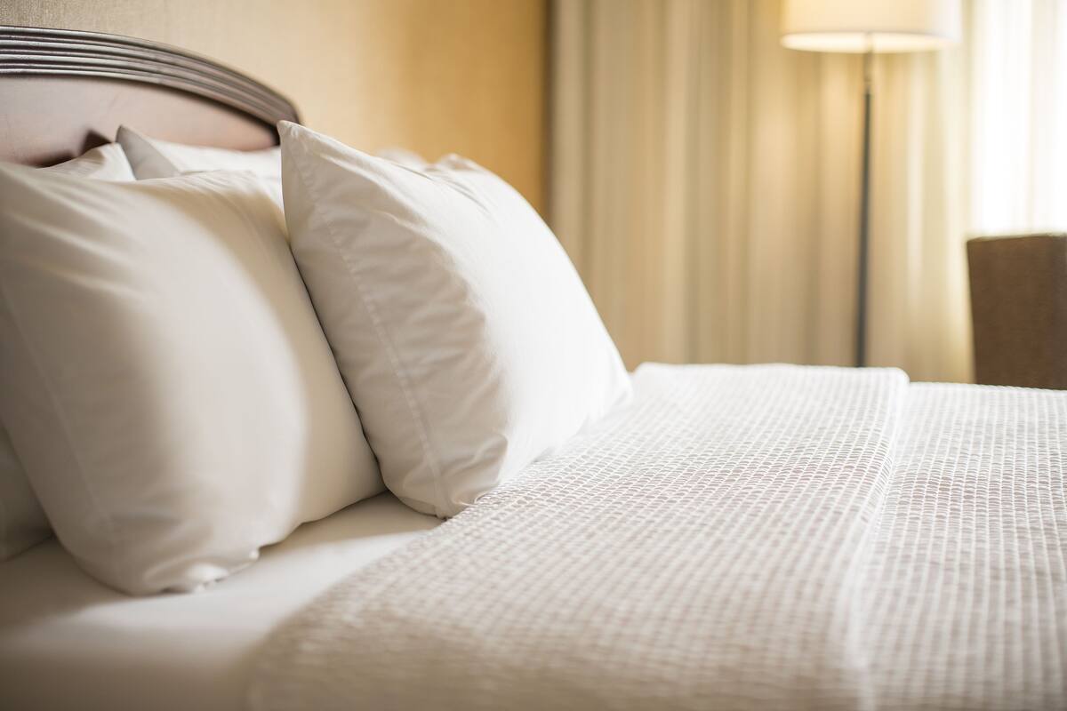 A neatly made bed with white bedding and pillows, a headboard, and a lamp in the background.