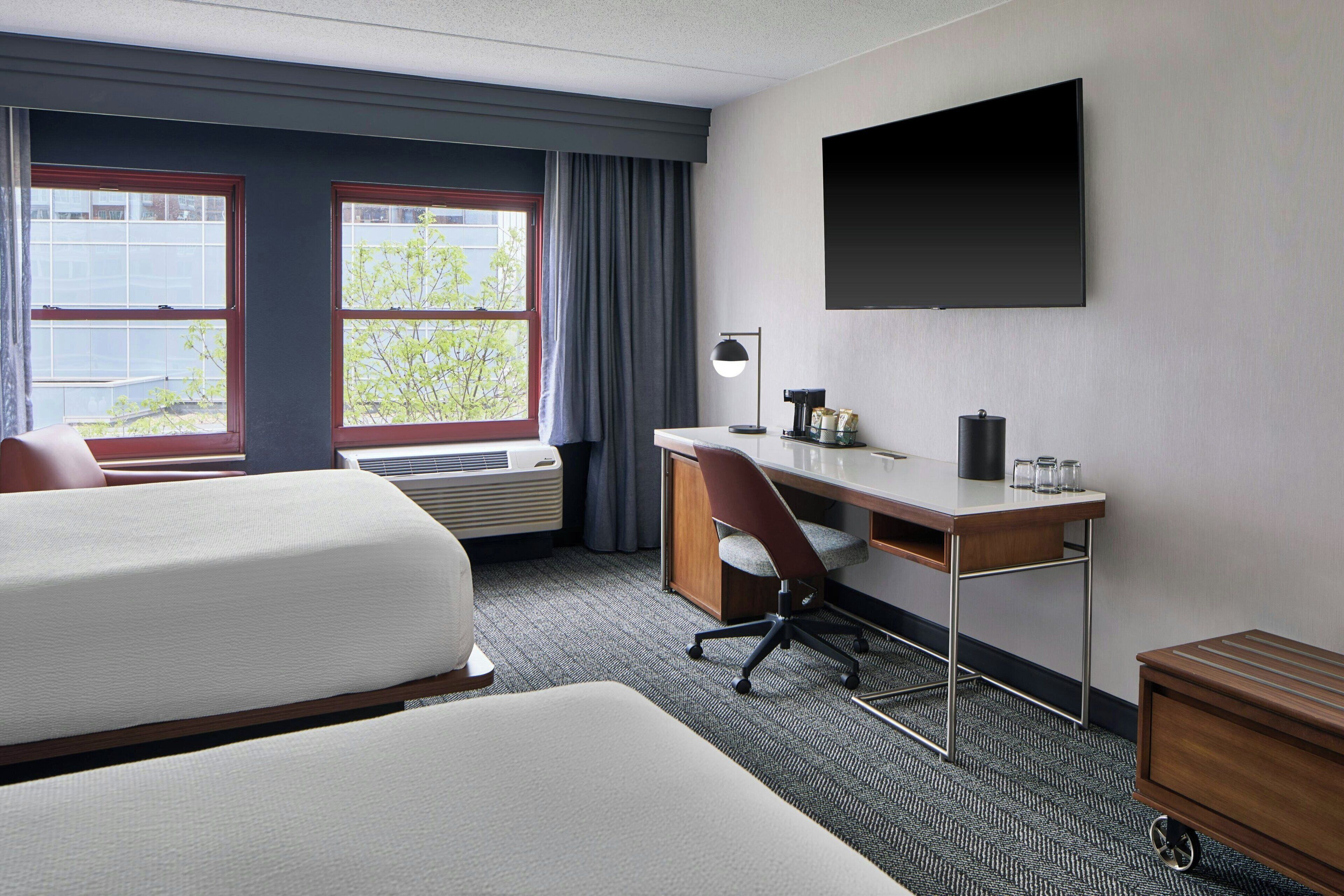 A hotel room with two beds, a desk, a chair, a TV, and a window with curtains.
