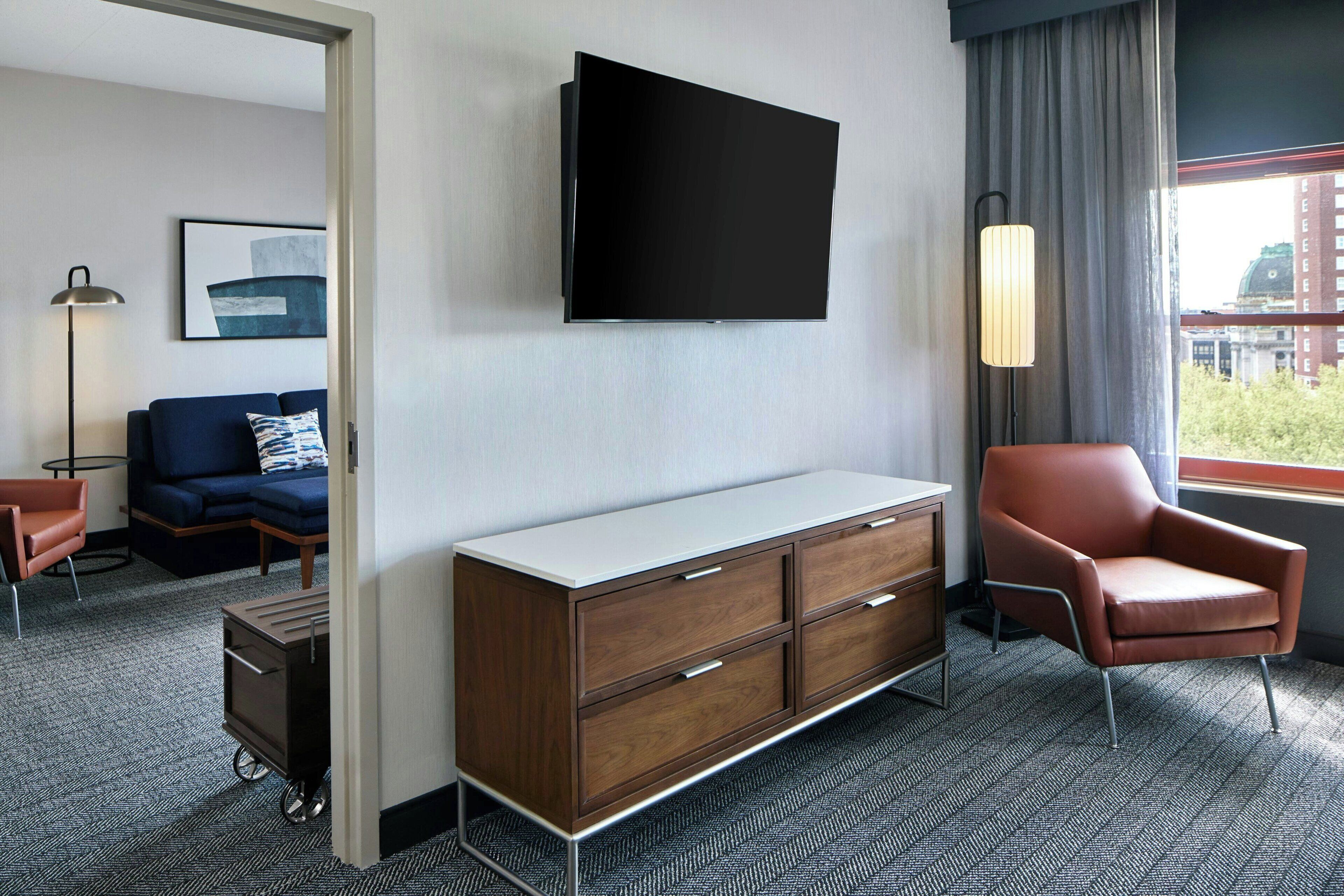 Quarto de hotel com televisão de tela plana, sofá, poltrona, armário e vista para a cidade.