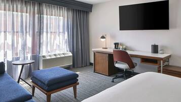 A hotel room with a bed, a desk, a chair, a small table, a TV, and a window with curtains.