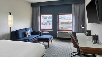 A hotel room with a bed, a sofa, a desk, a chair, a lamp, a TV, and a window with curtains.