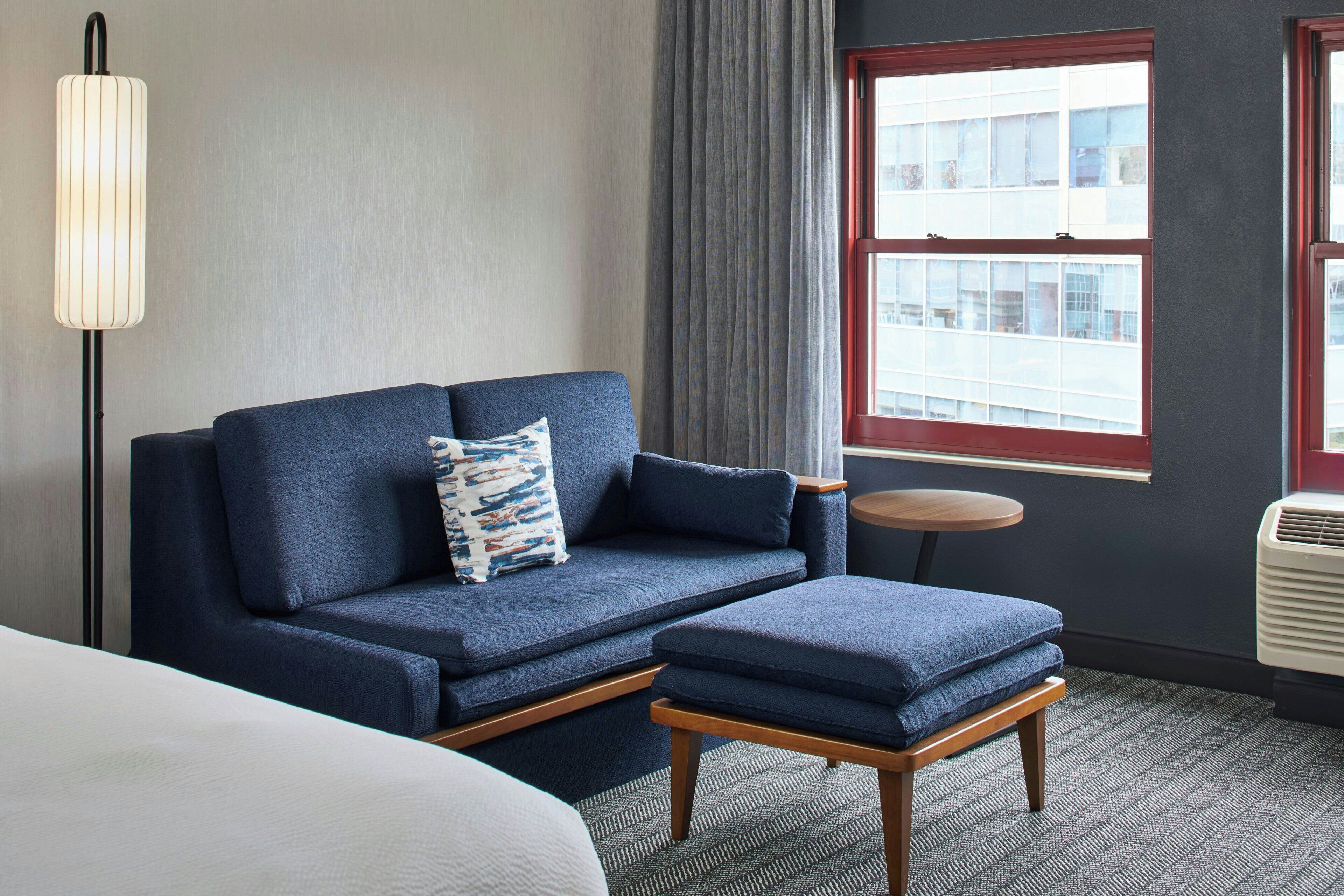 A room with a sofa, a small table, a lamp, a window, and an air conditioning unit.