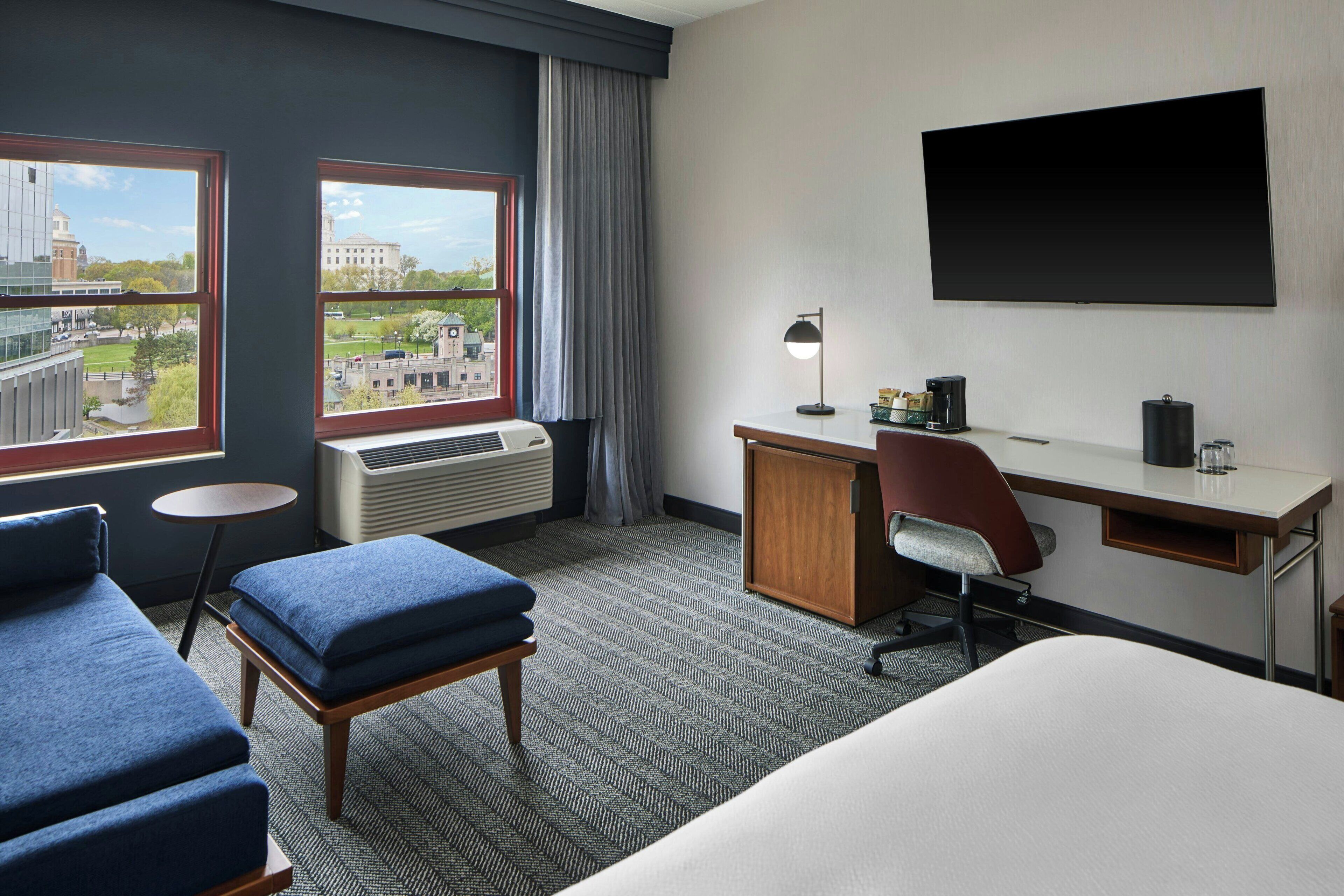A hotel room with a bed, a desk, a chair, a small table, and a TV.