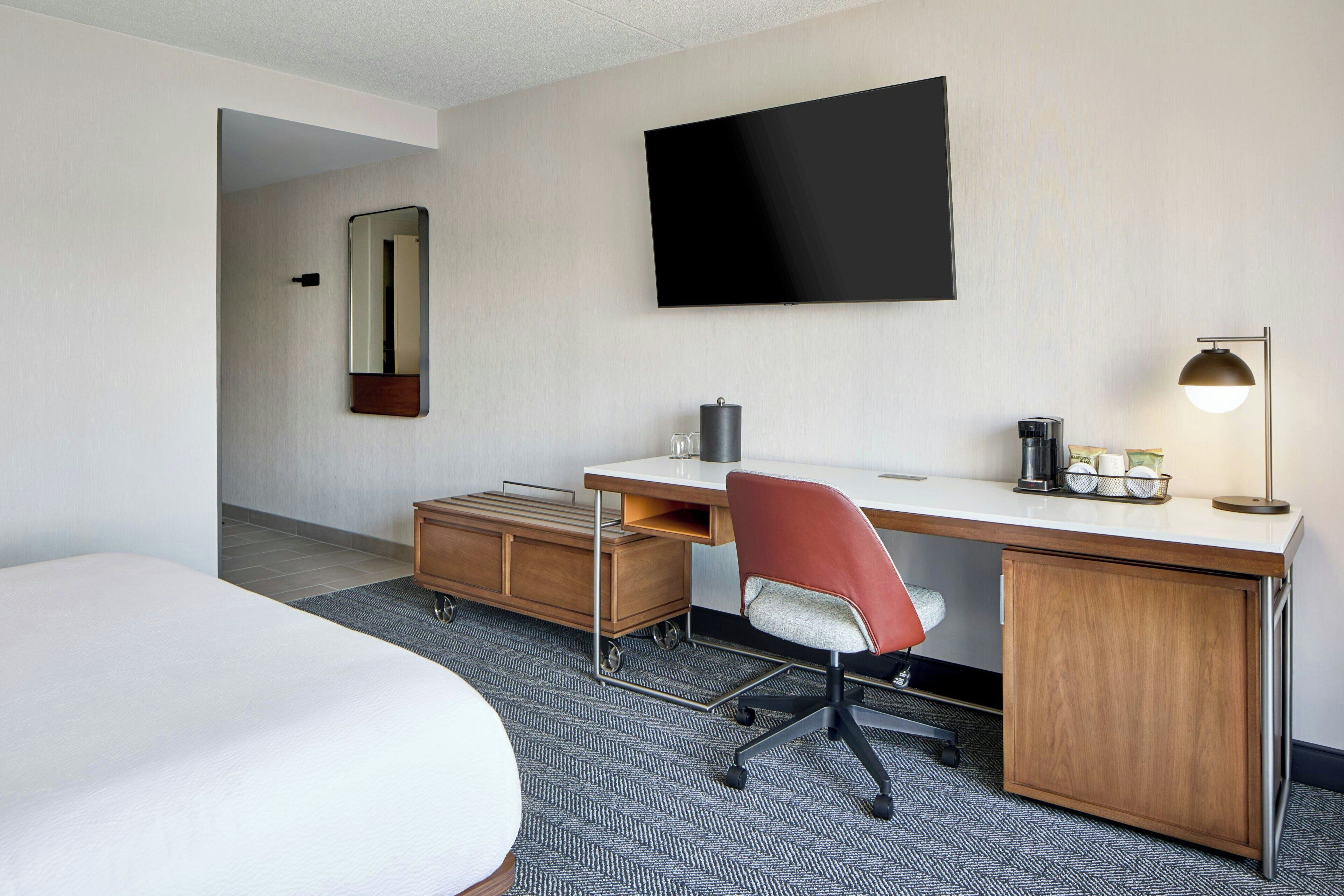 Una habitación de hotel con una cama, un escritorio con silla, televisor, lámpara y cafetera.