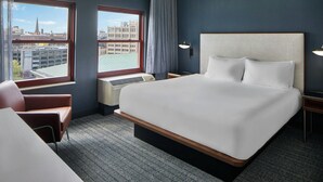 A hotel room with a large bed, a chair, a desk, a window with a view of buildings, and a wall-mounted lamp. - Courtyard by Marriott Providence Downtown (Providence)