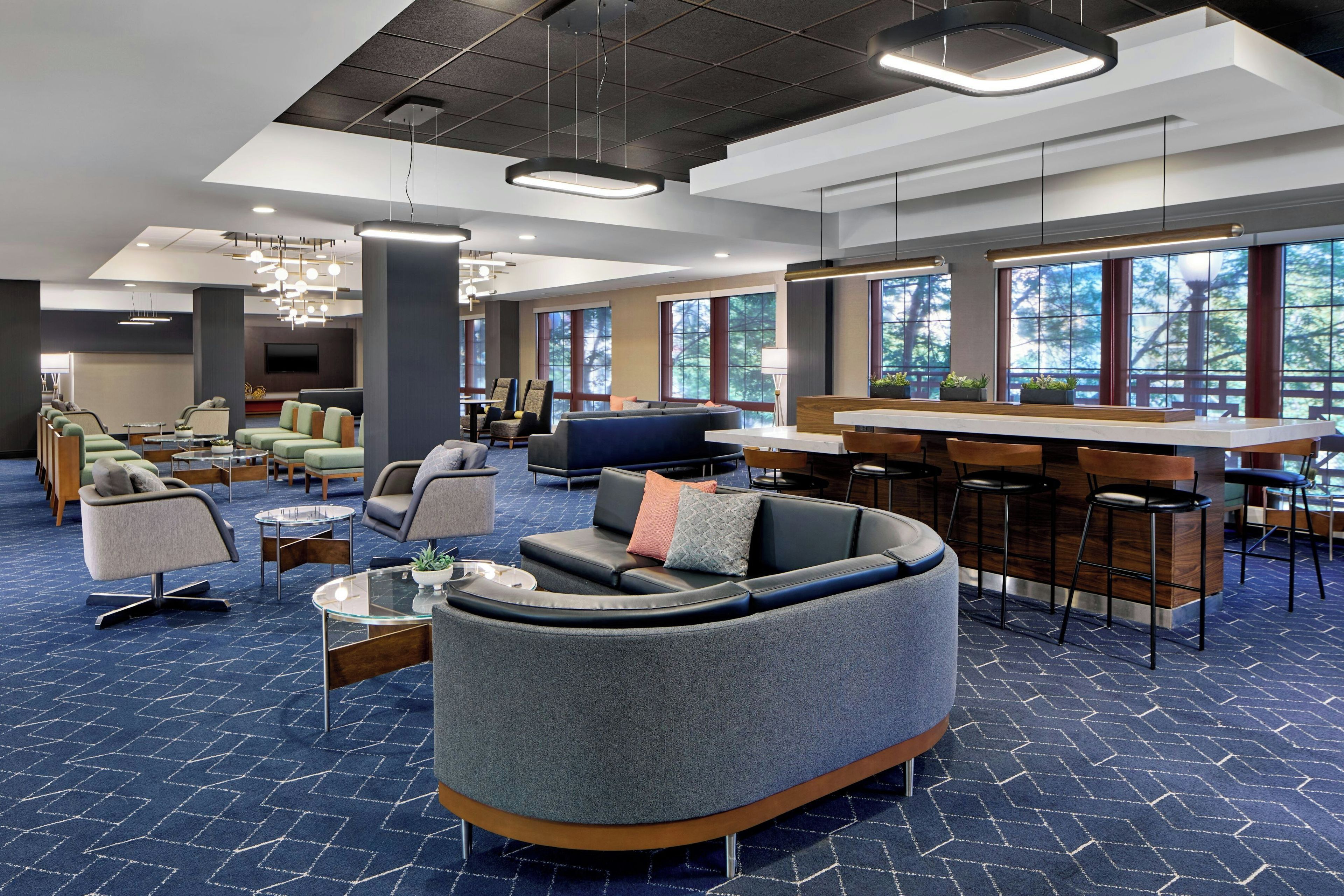 A modern hotel lobby with seating areas, a bar, and large windows offering views of trees.