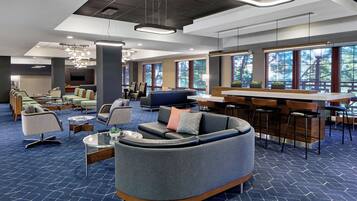 A modern hotel lobby with seating areas, a bar, and large windows offering views of trees.