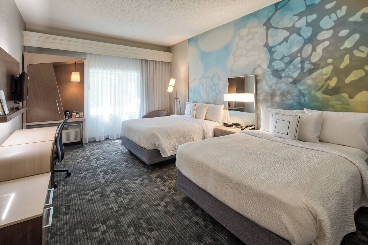 Ein Hotelzimmer mit zwei Betten, einem Schreibtisch, einem Sessel, einem Fernseher und einem großen, abstrakten Wandbild.