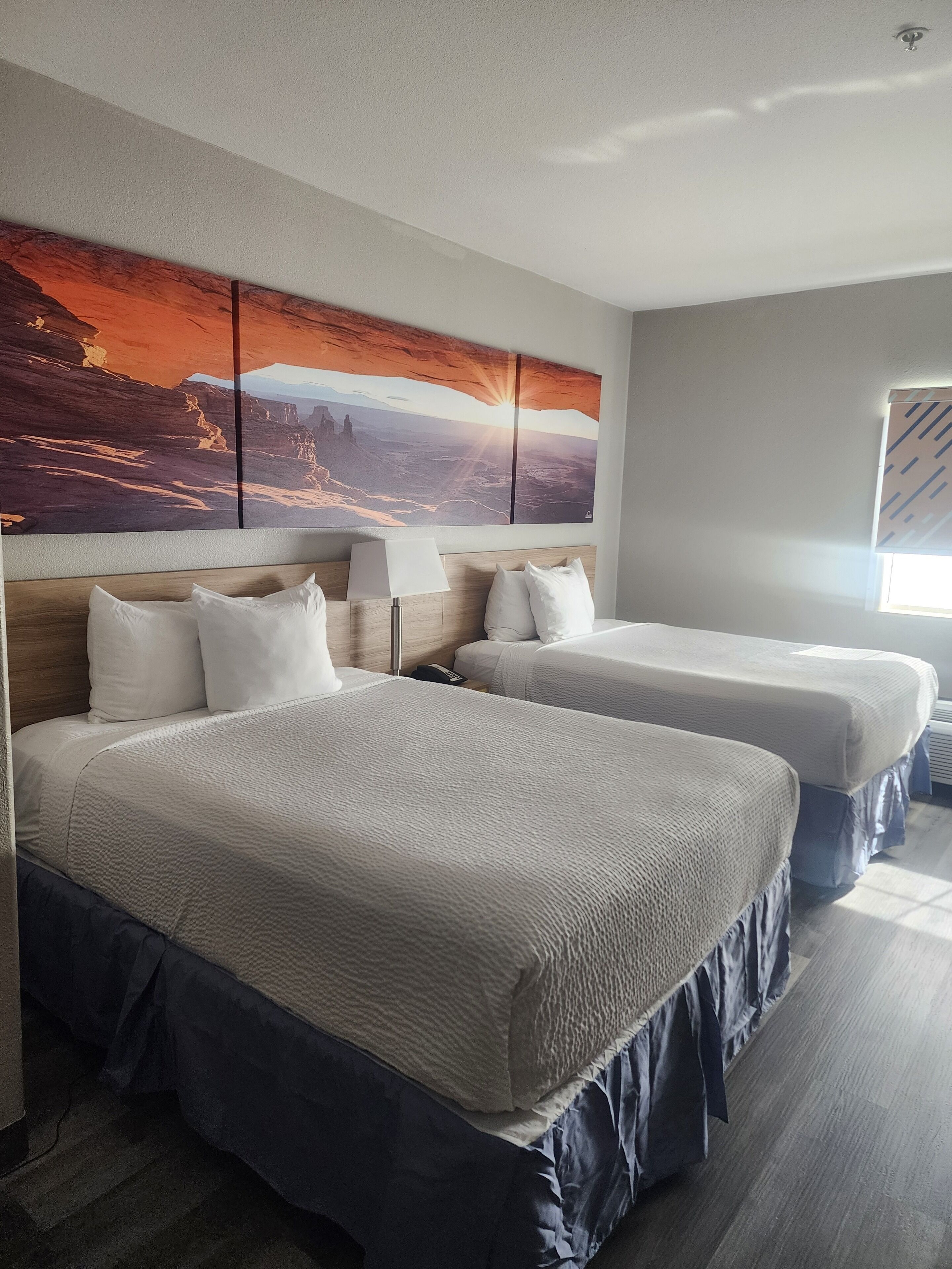 Habitación de hotel con dos camas, un cuadro grande en la pared, una ventana con persianas y una lámpara en el techo.