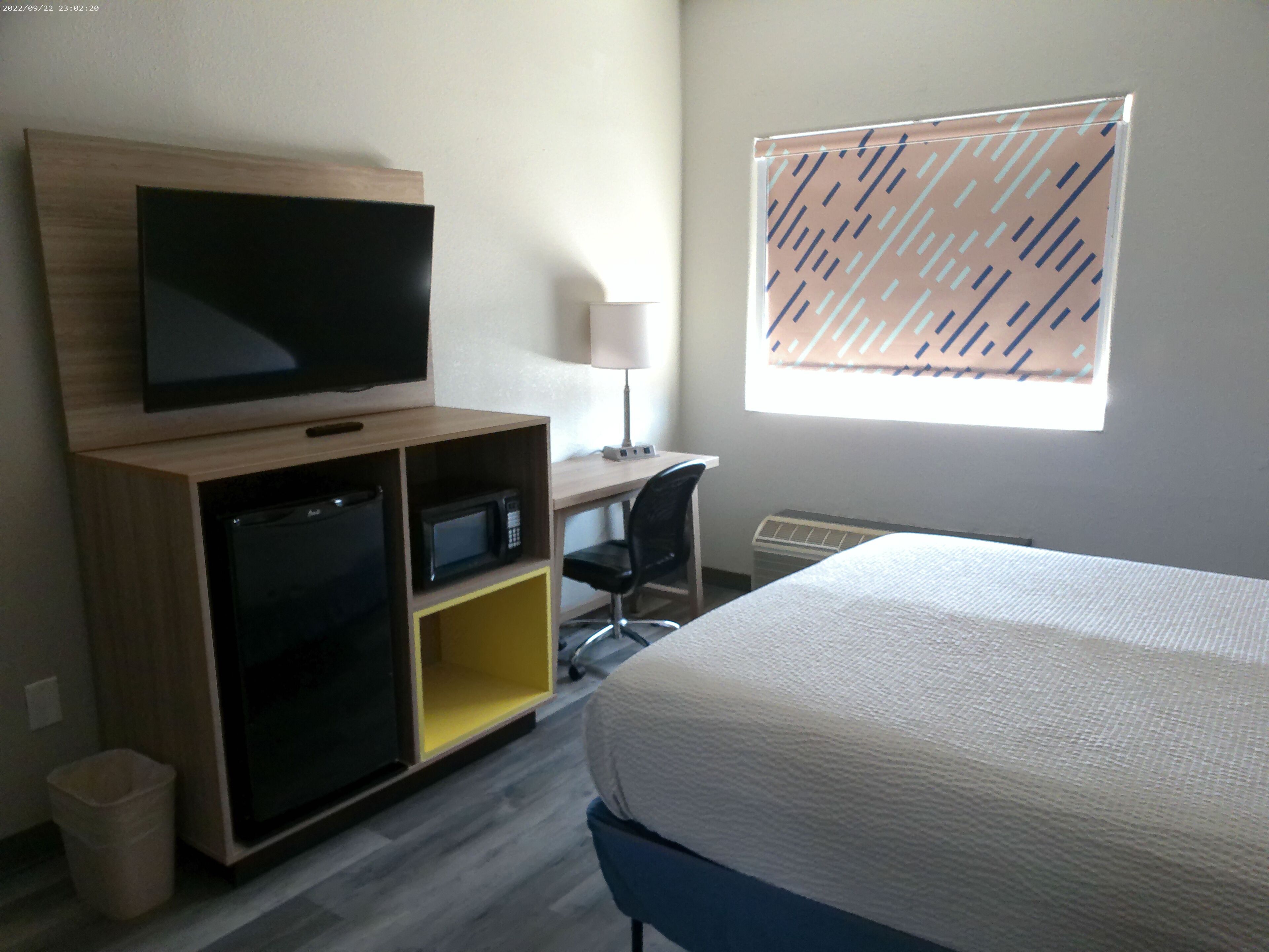 Habitación de hotel con cama, televisión, escritorio con silla, microondas y ventana con persianas.