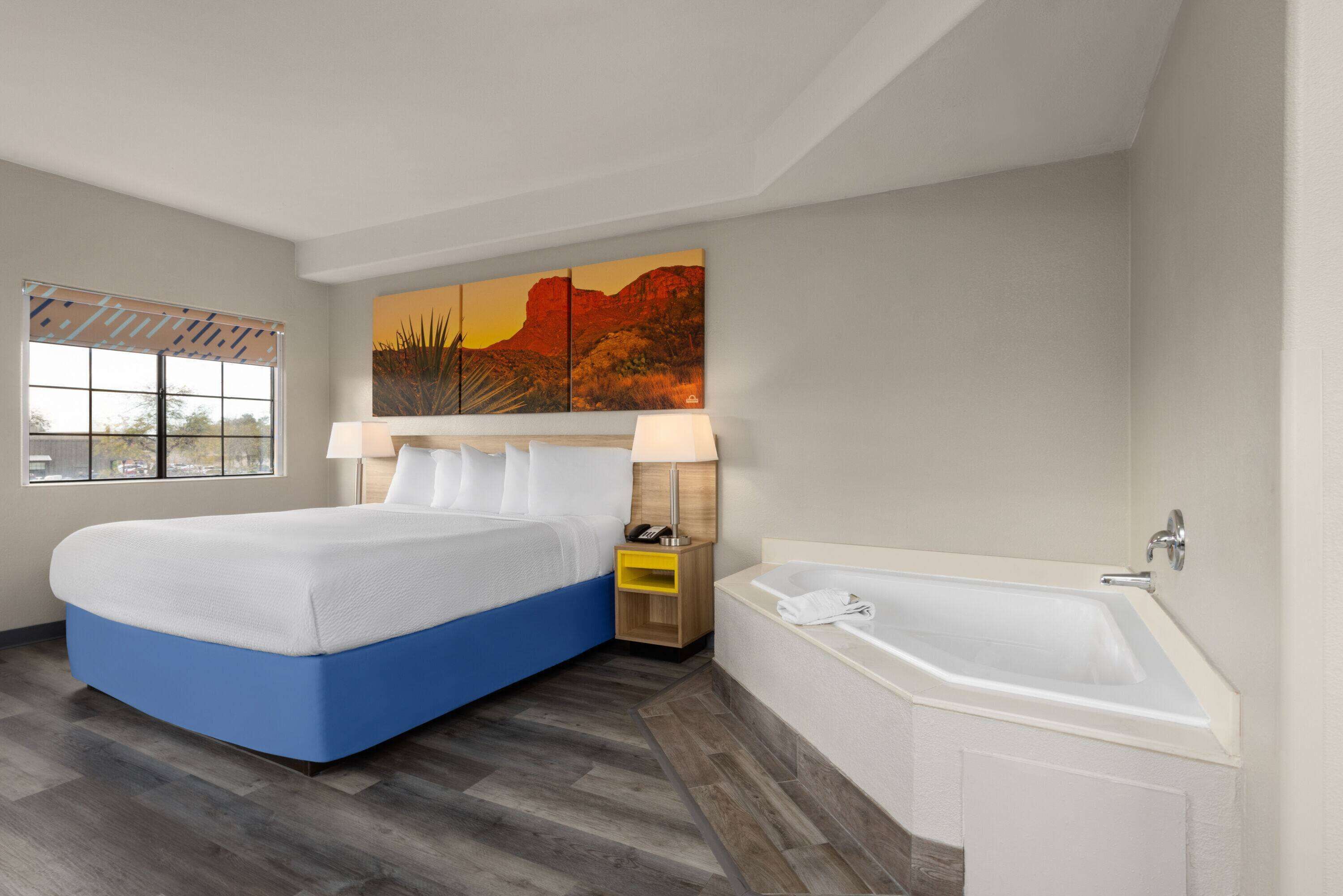 A hotel room with a large bed, a bathtub, a window with blinds, and a painting on the wall.