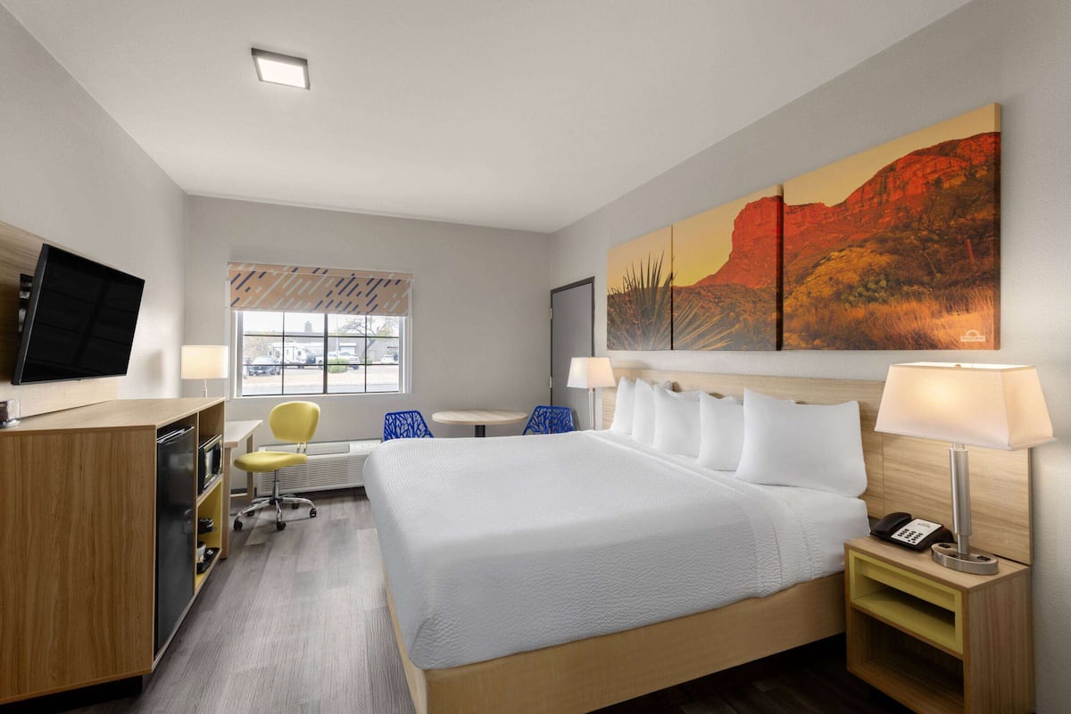 Habitación de hotel con una cama grande, un escritorio con computadora, una silla, una ventana con persianas y un cuadro en la pared.