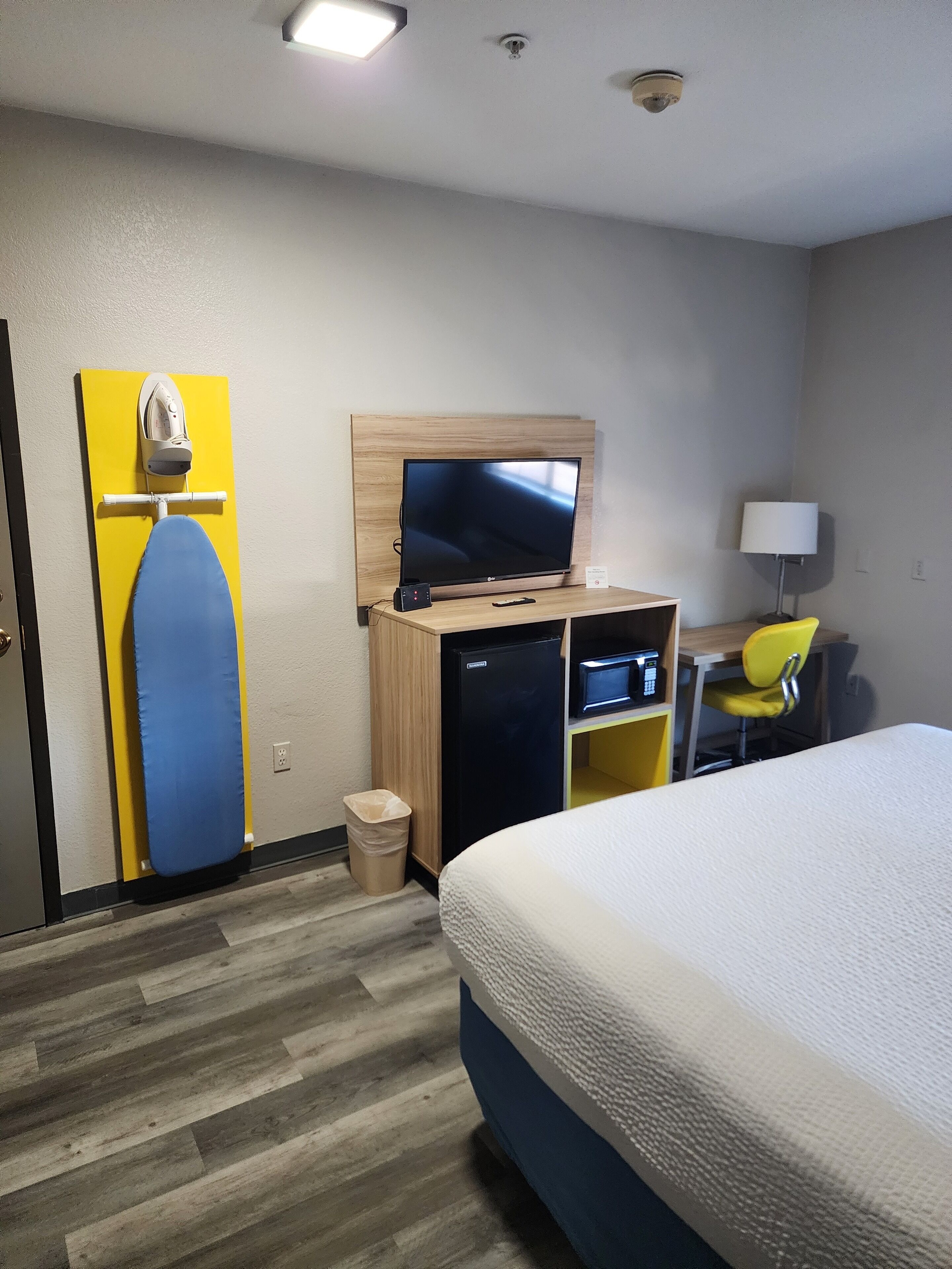 A hotel room with a bed, a desk with a computer, a TV, a chair, and a shoe rack.