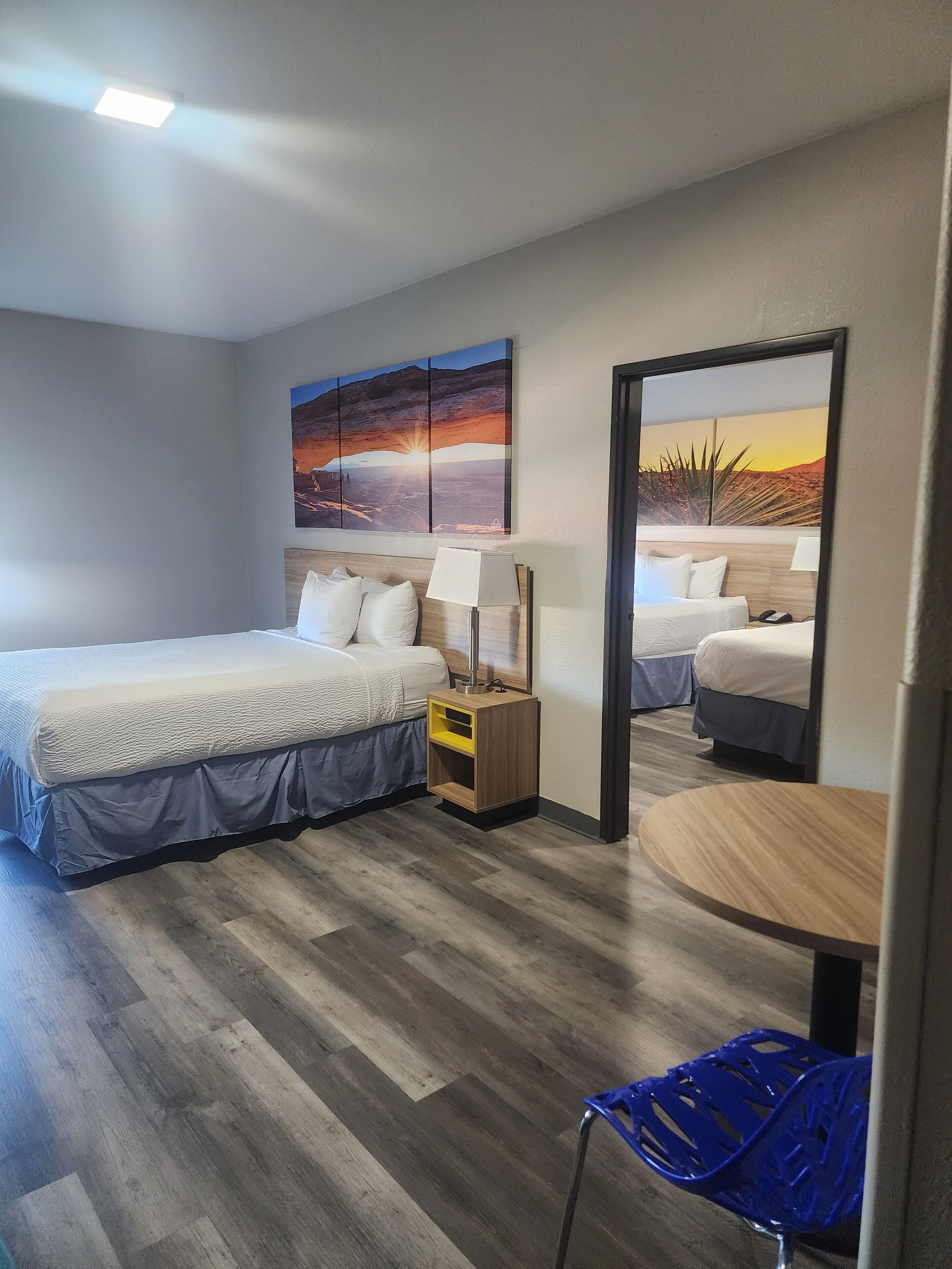 Een hotelkamer met twee bedden, een kleine tafel, een stoel, een nachtkastje en een schilderij aan de muur.