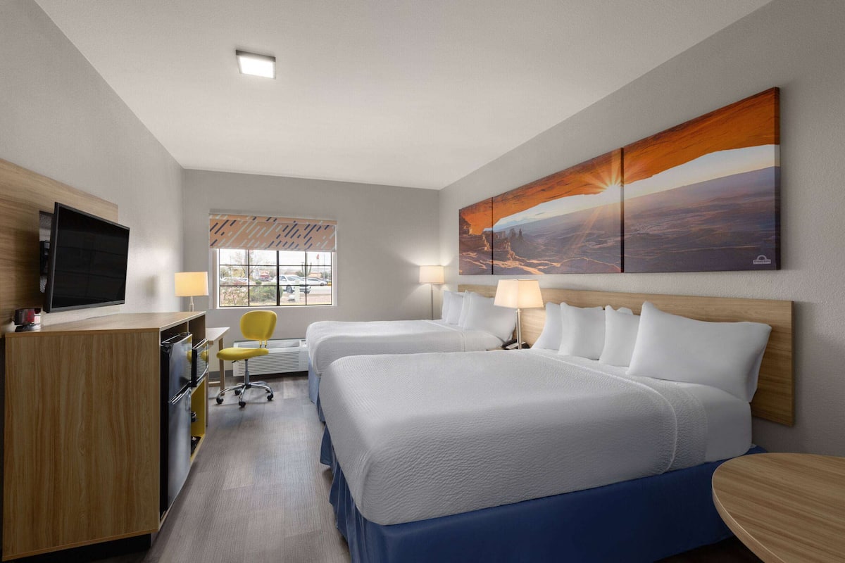 Una habitación de hotel con dos camas, un escritorio con televisor, una silla y una ventana con persianas.