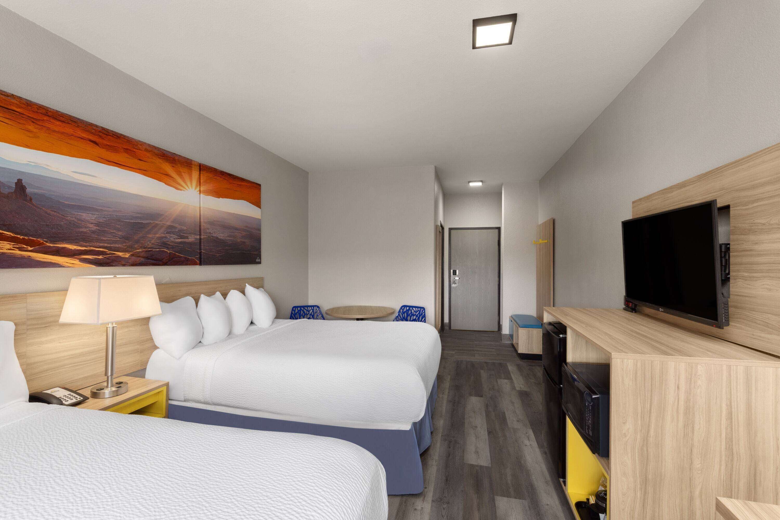 A hotel room with two beds, a TV, a desk, a lamp, and a painting of a sunset.