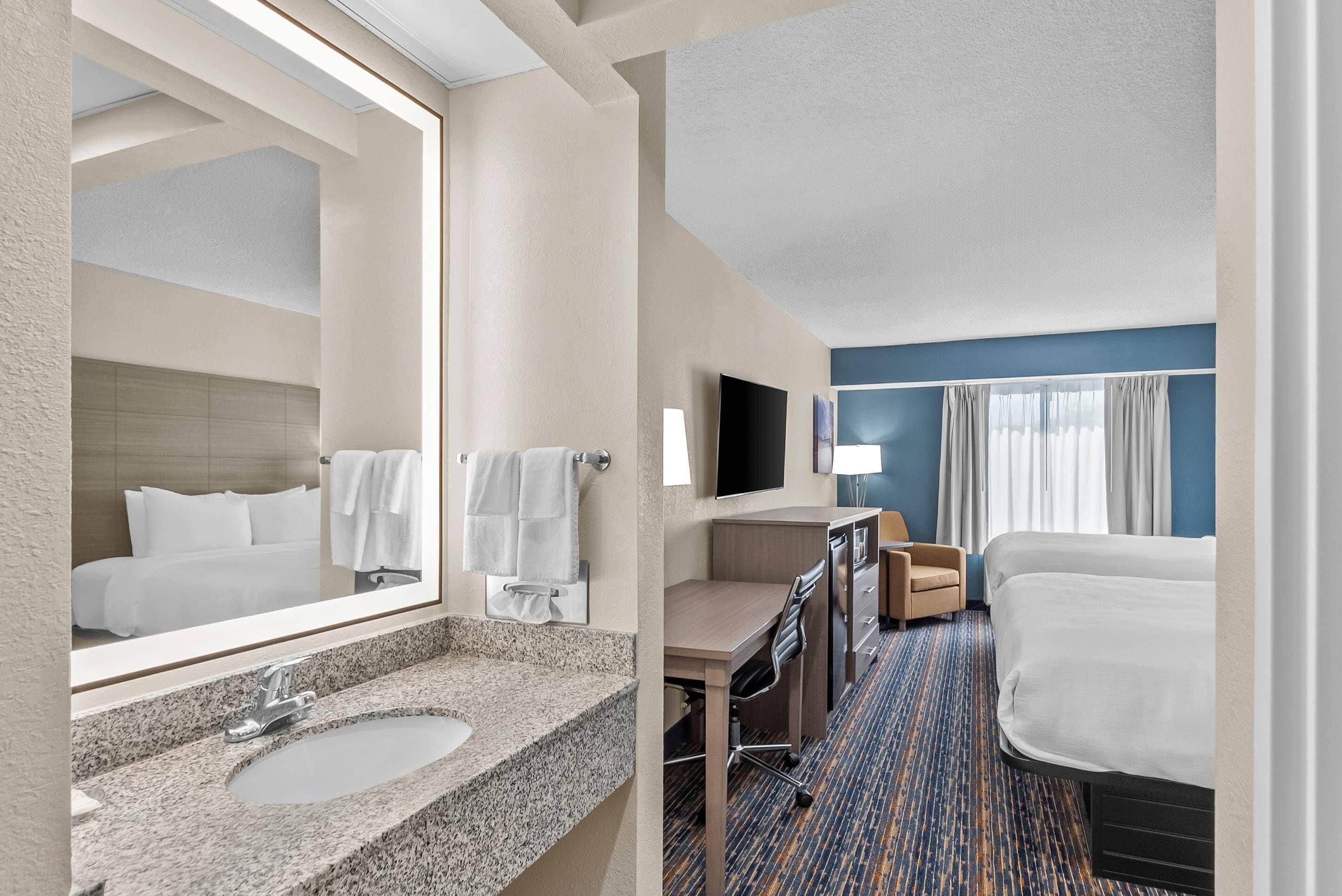 A hotel room with a large mirror, a sink, a desk, a chair, a flat-screen TV, a bed, and a window with curtains.