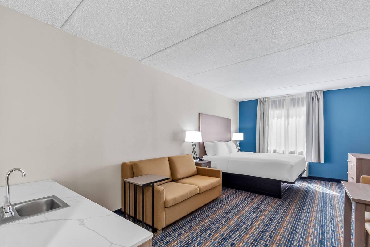 A hotel room with a bed, a sofa, a desk, a lamp, a nightstand, and a bathroom with a sink.