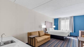 A hotel room with a bed, a sofa, a desk, a lamp, a nightstand, and a bathroom with a sink.