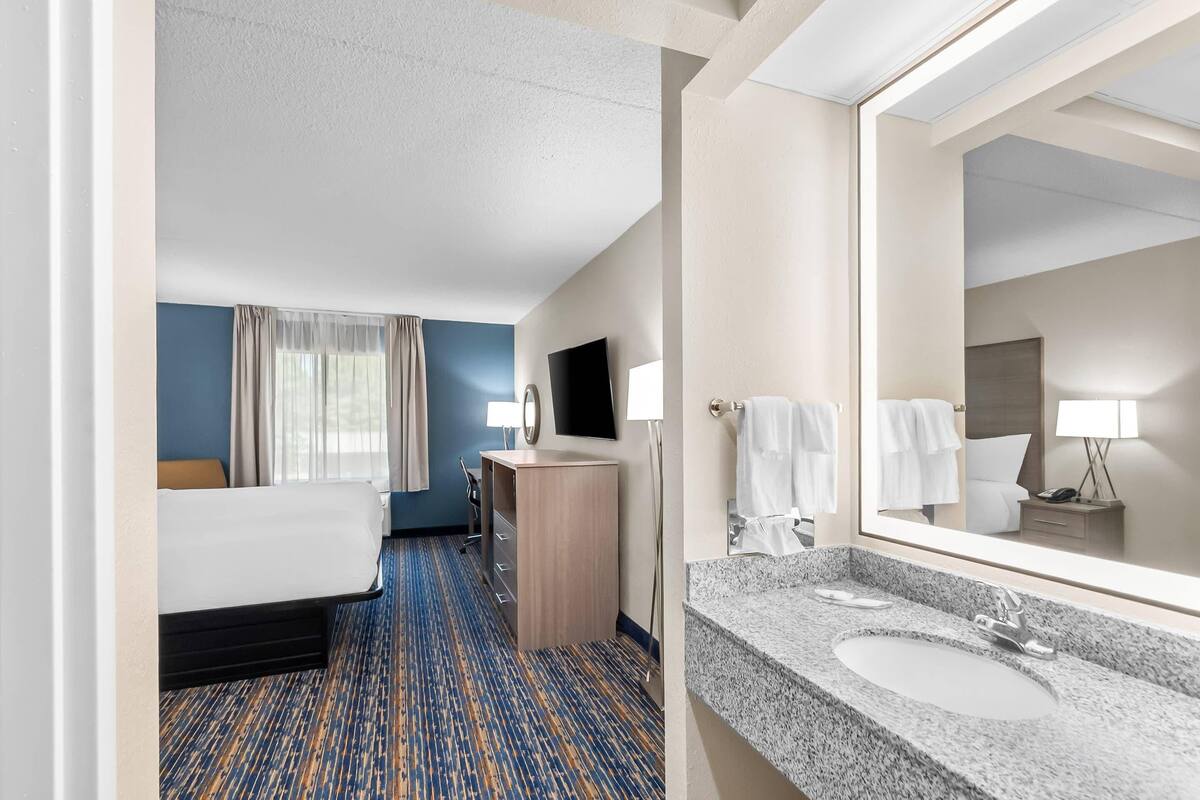 A hotel room with a bed, a desk, a chair, a desk lamp, a flat-screen TV, a bathroom with a sink and mirror, and a patterned carpet.